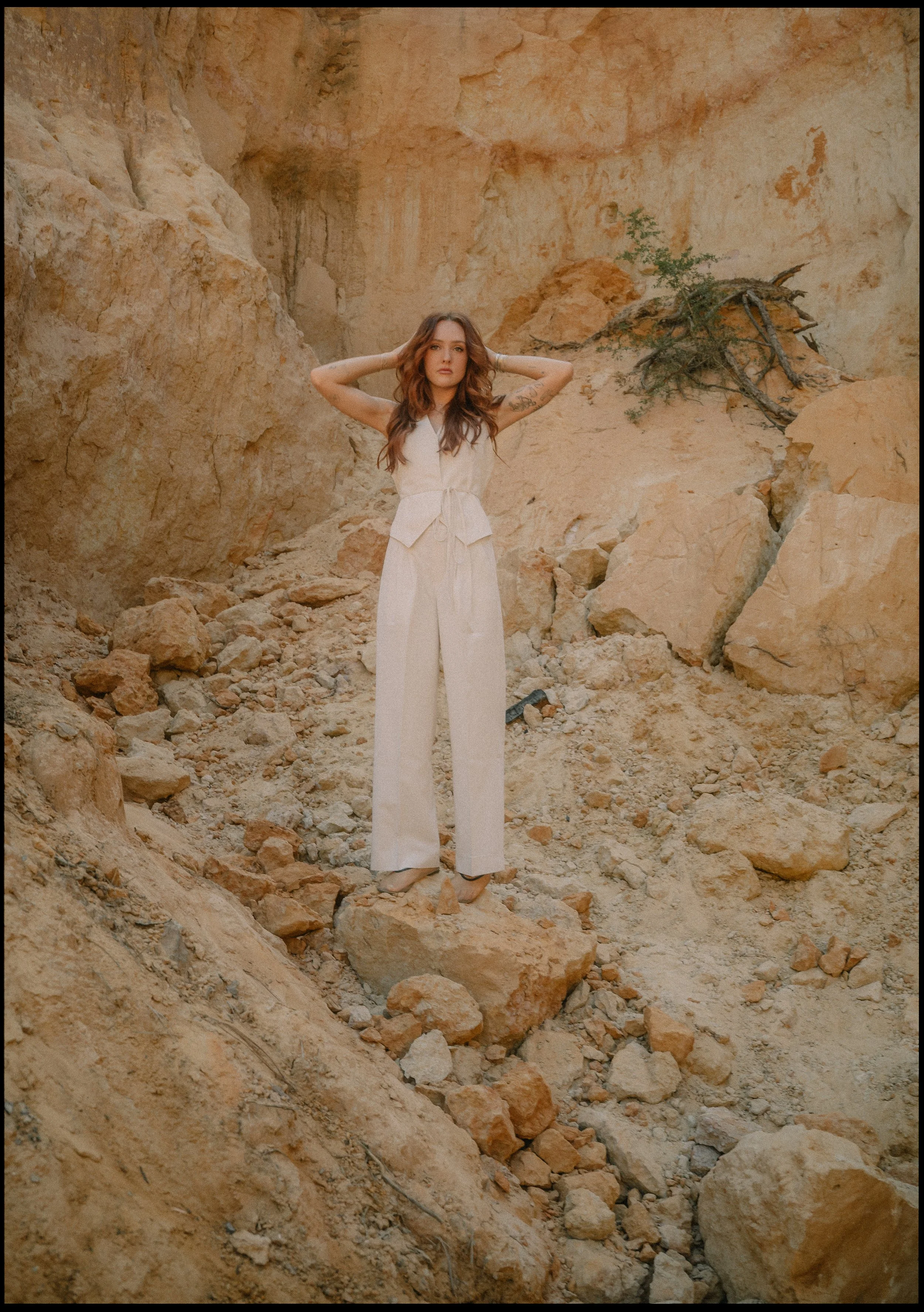 A woman wearing a white jumpsuit standing on rocks in a desert landscape with rocky cliffs and sparse vegetation.