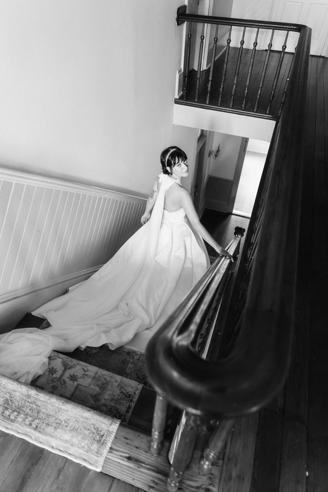 A woman in a wedding dress with a train descending stairs inside a house.