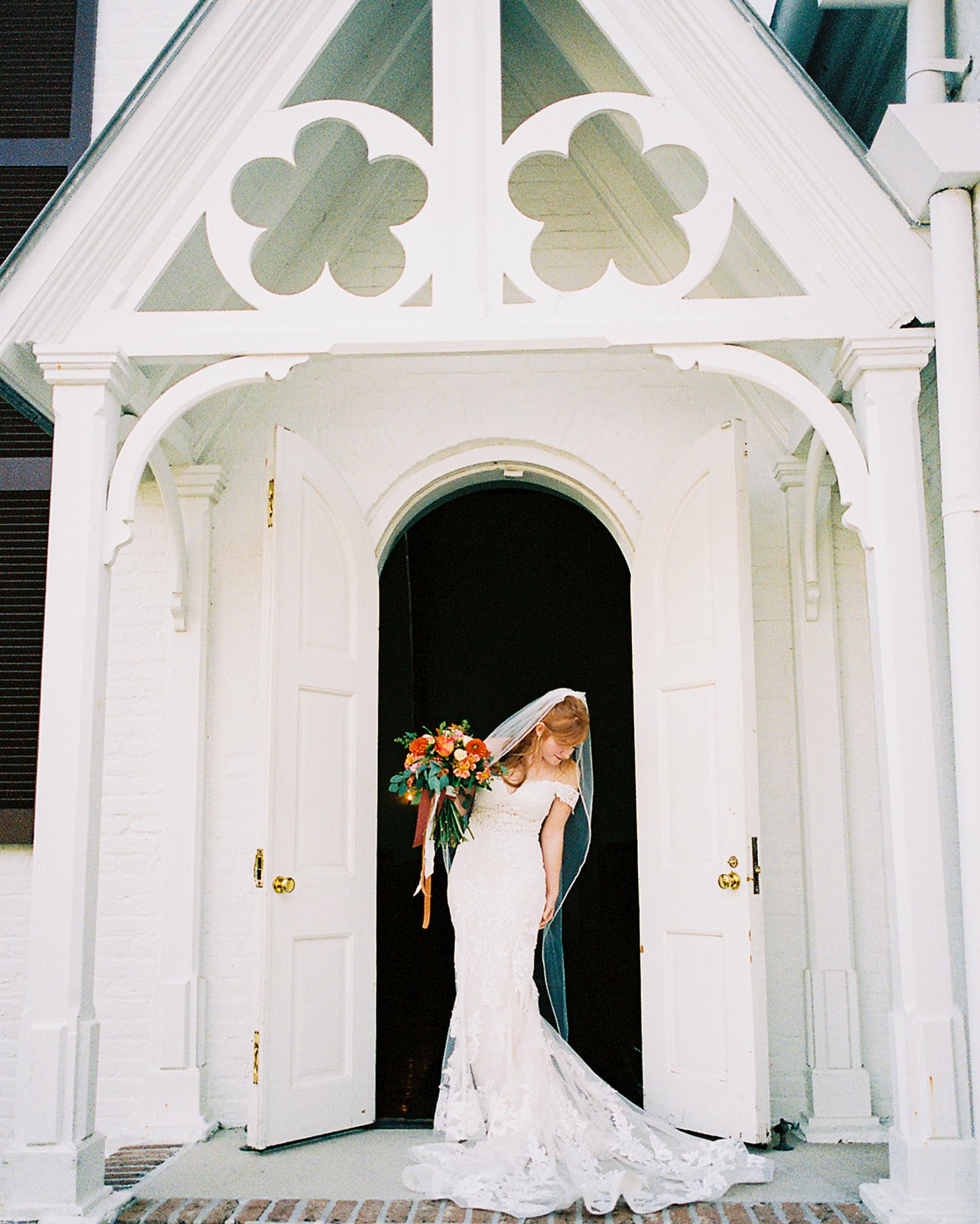 A bride in a white lace wedding dress standing at the doorway of a white house with open double doors, holding a colorful bouquet of flowers and looking downward.