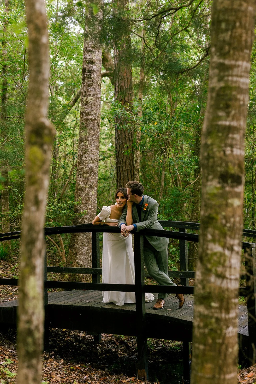 A couple is on a small wooden bridge surrounded by tall trees and dense green foliage, sharing a moment of intimacy.