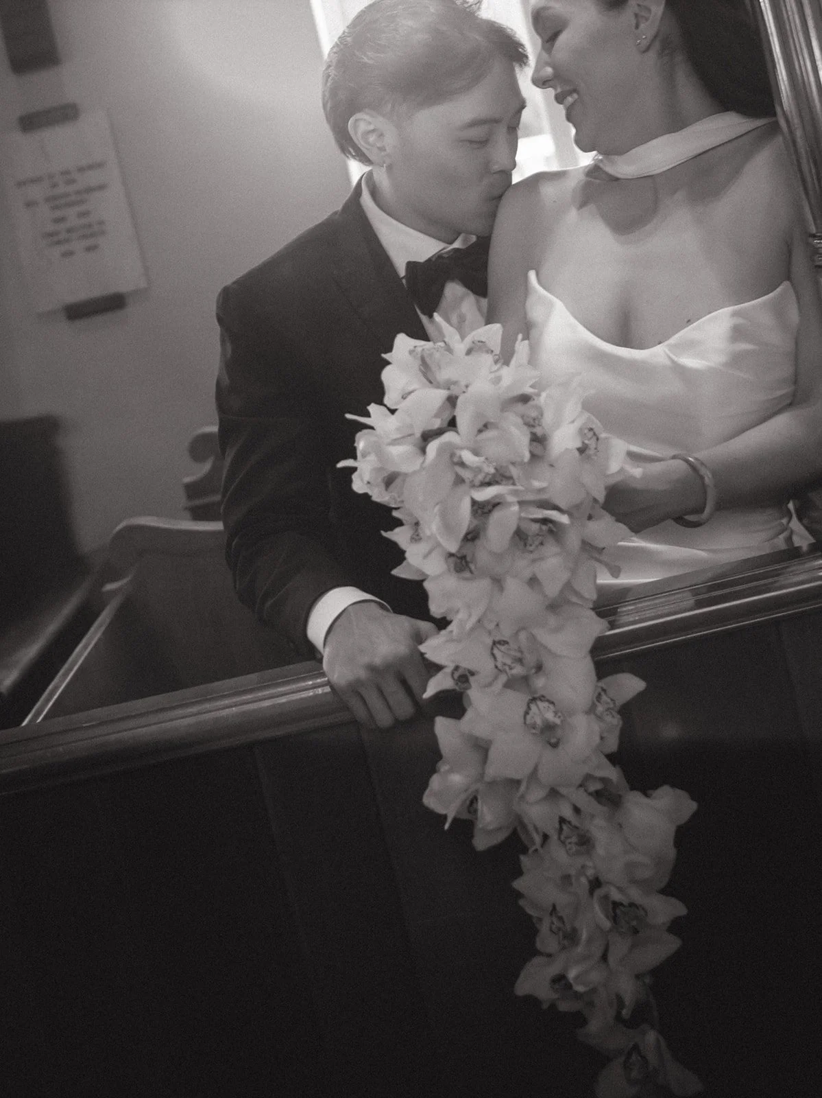 Black and white photo of a groom in a tuxedo kissing a bride in a wedding dress on the cheek, both smiling, with the bride holding a cascading bouquet of flowers.