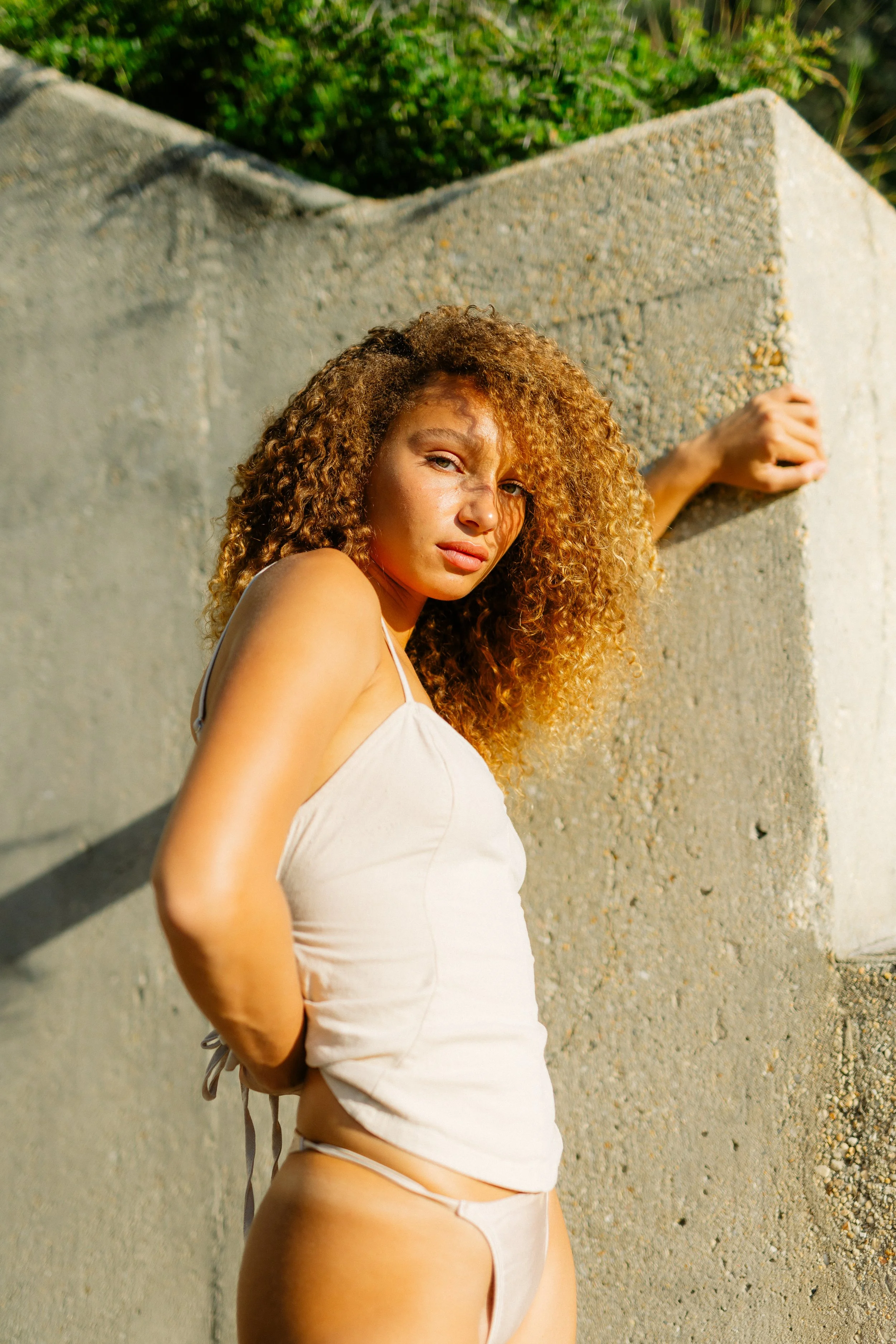 A woman with curly hair posing beside a concrete wall outdoors during sunset.