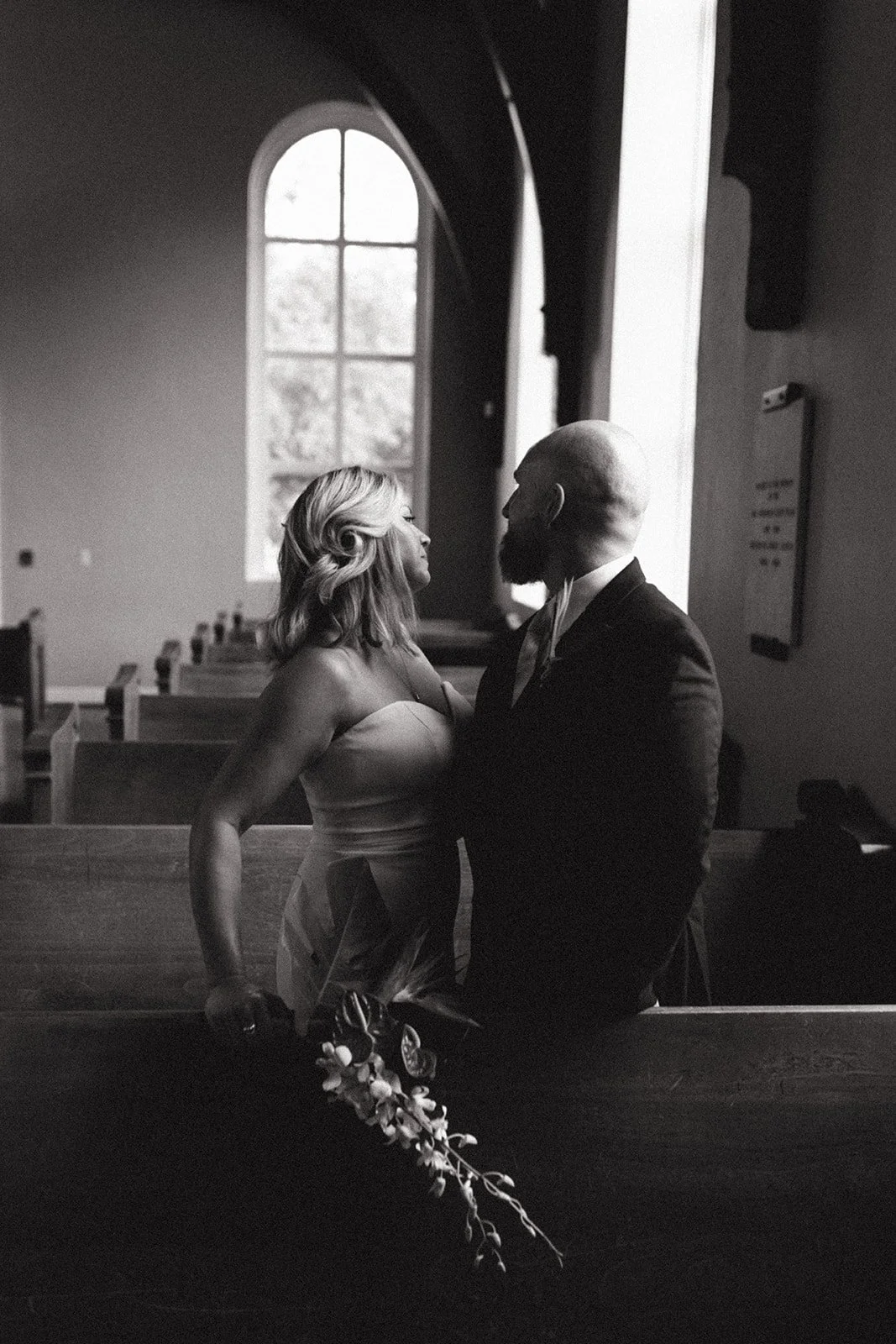 A black and white photo of a couple inside a church, standing close and looking into each other's eyes, with a large arched window behind them.