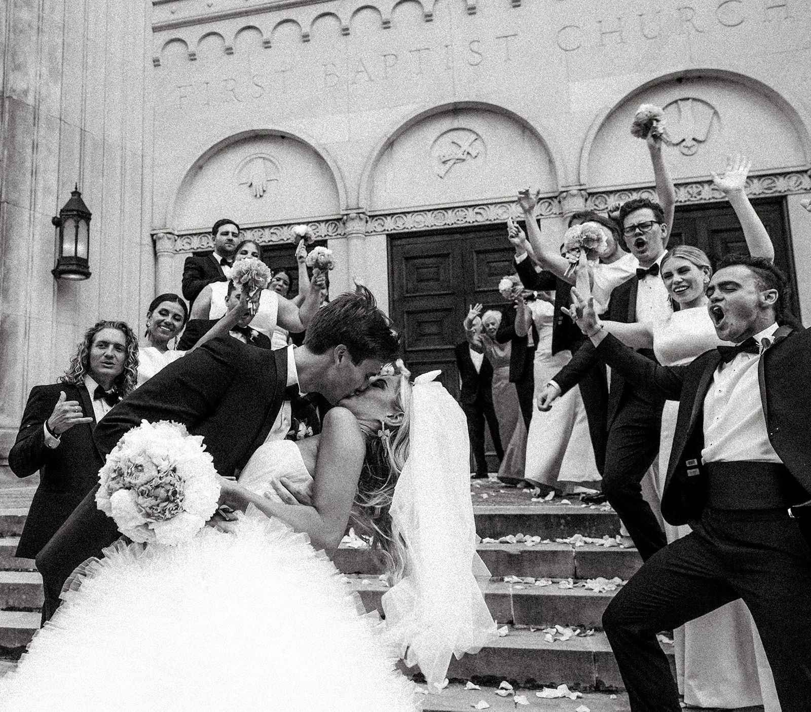 A wedding celebration with a bride and groom kissing on church steps surrounded by friends and family. The guests are cheering and celebrating, some waving bouquets and others singing.