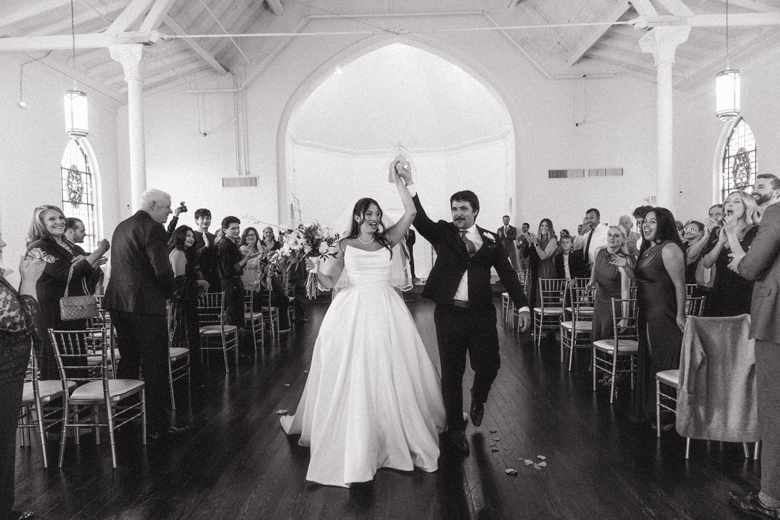 A bride and groom are walking down the aisle holding hands with their arms raised, smiling. The bride is wearing a white wedding gown, and the groom is in a dark suit. They are surrounded by friends and family inside a church with high ceilings and s