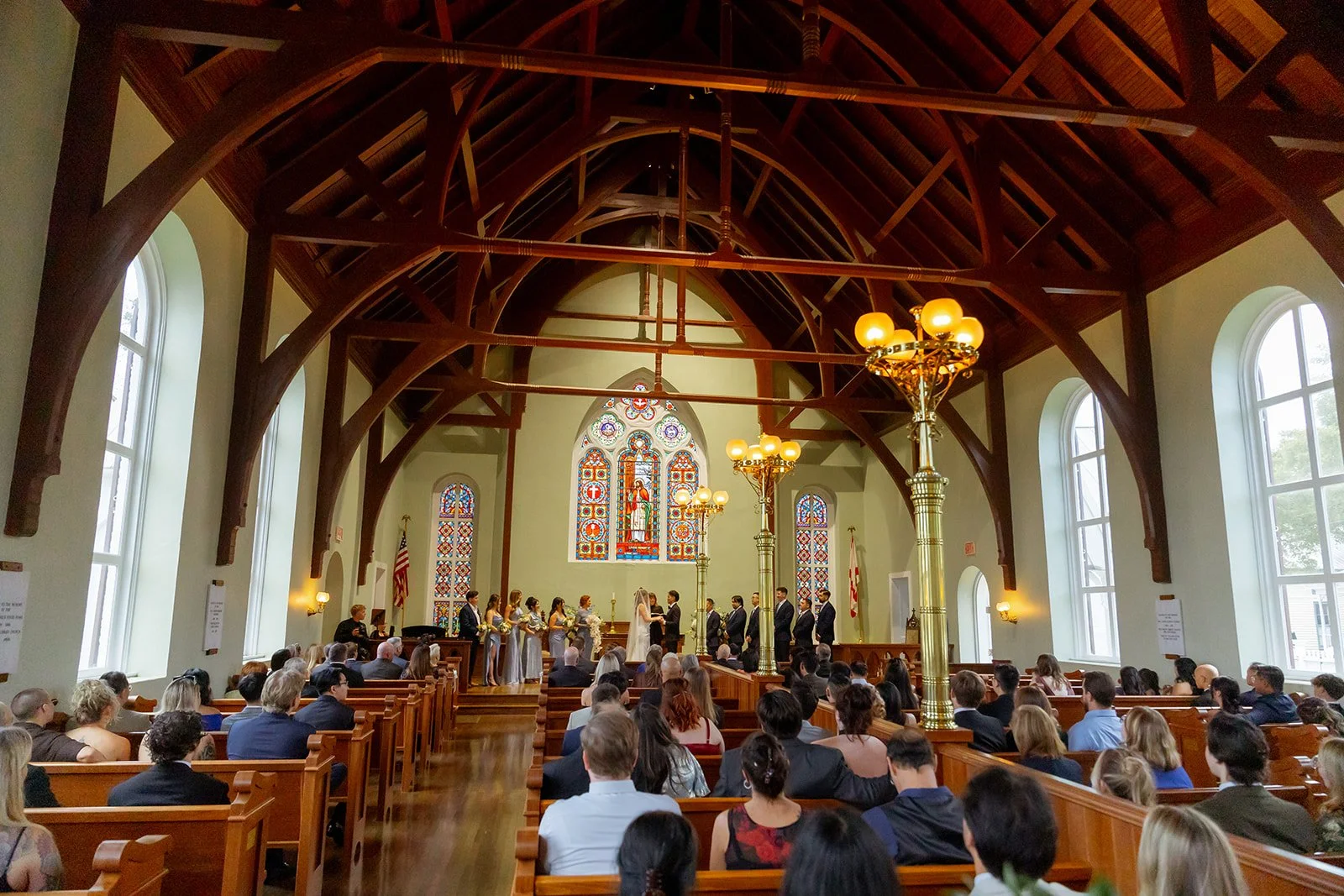 A wedding ceremony in a church with high wooden vaulted ceilings, stained glass windows, and a large congregation seated in pews, as the couple exchanges vows at the altar.