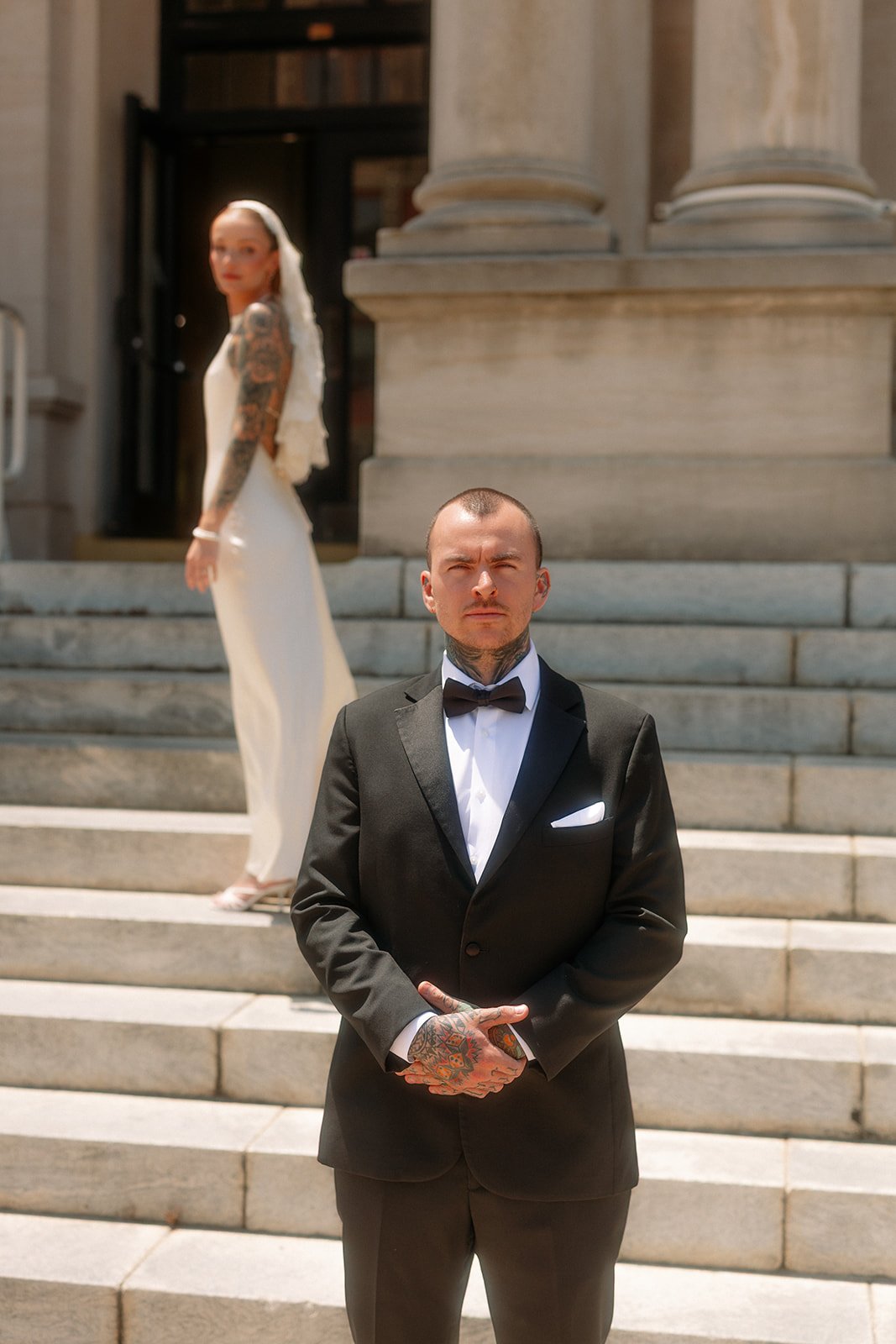 A man in a tuxedo with a bow tie and tattoos on his hands stands in front of a building with stone stairs. A woman in a white dress and tattoos on her arm stands on the stairs behind him.