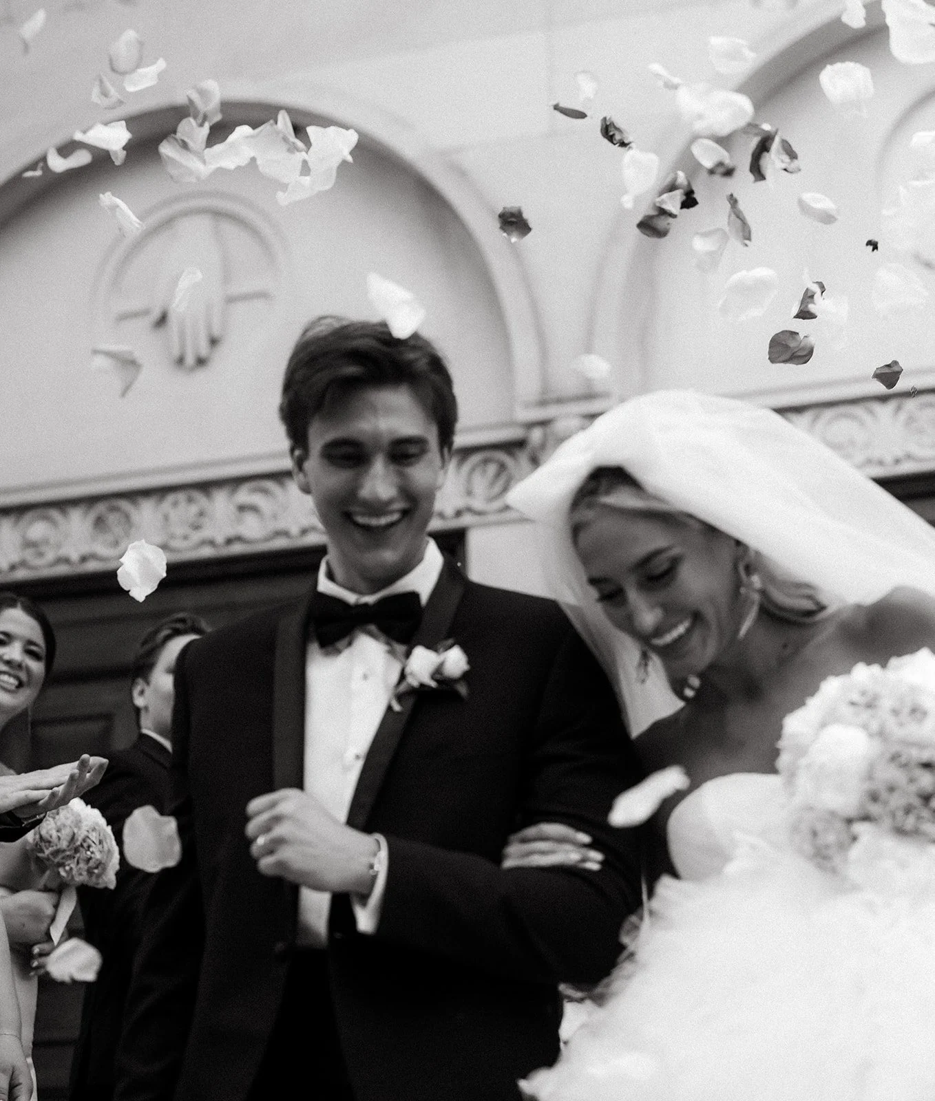 A happy newlywed couple at their wedding reception surrounded by friends, with rose petals falling around them. The groom is in a tuxedo and the bride is in a wedding dress with a veil, both smiling.