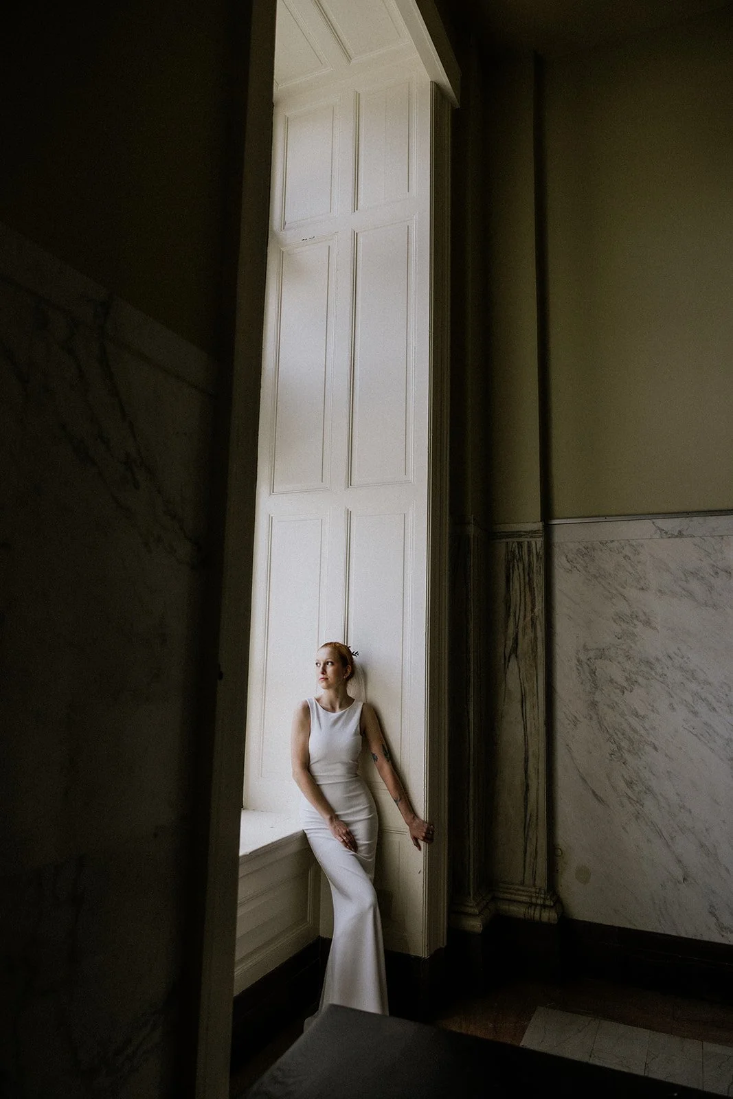 A woman in a white sleeveless dress standing by a large window with white paneling in an elegant room with marble walls.