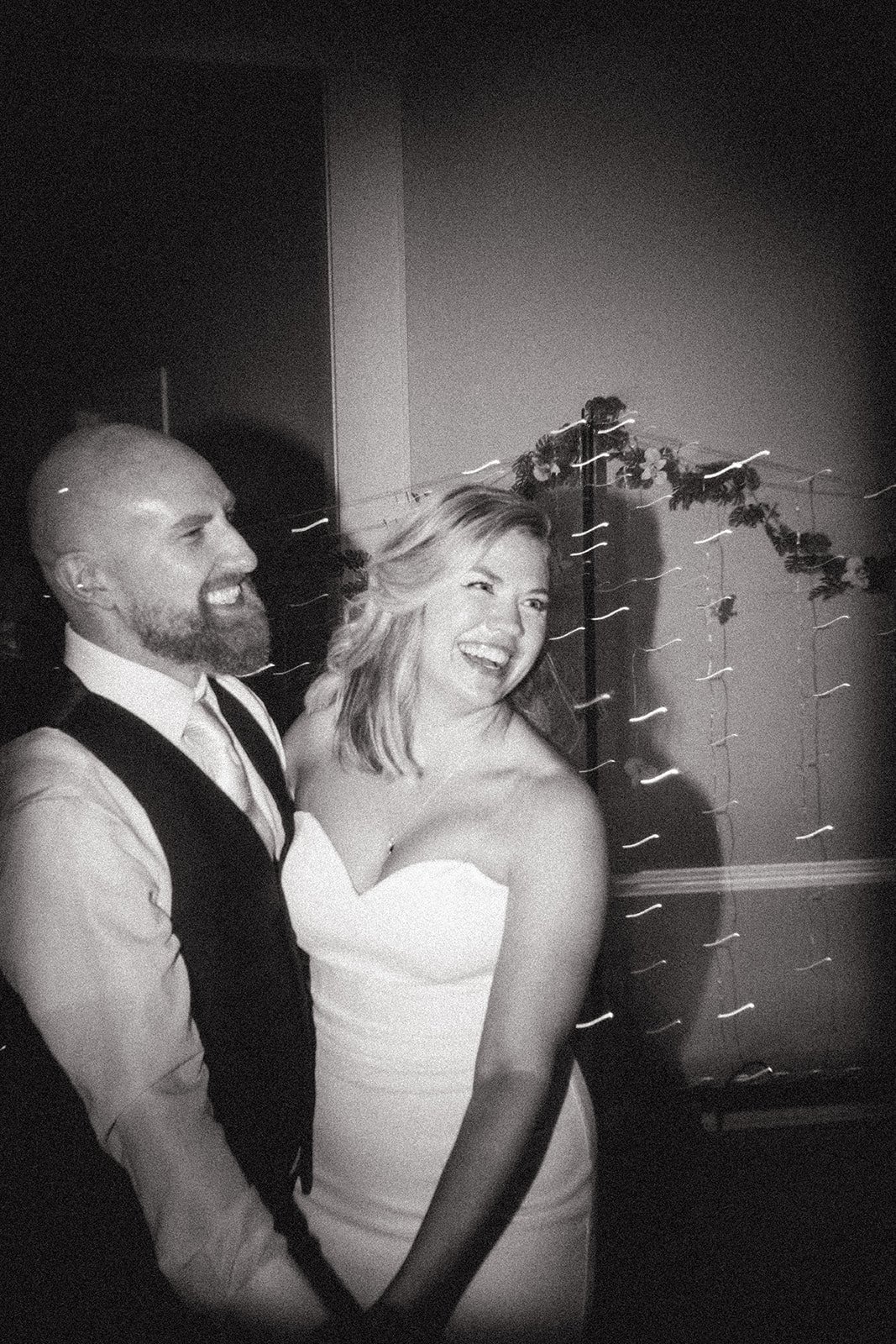 A black-and-white photo of a smiling woman in a strapless dress and a smiling man in a shirt and vest, standing close together at an indoor event, with blurred decorative lights and flowers in the background.