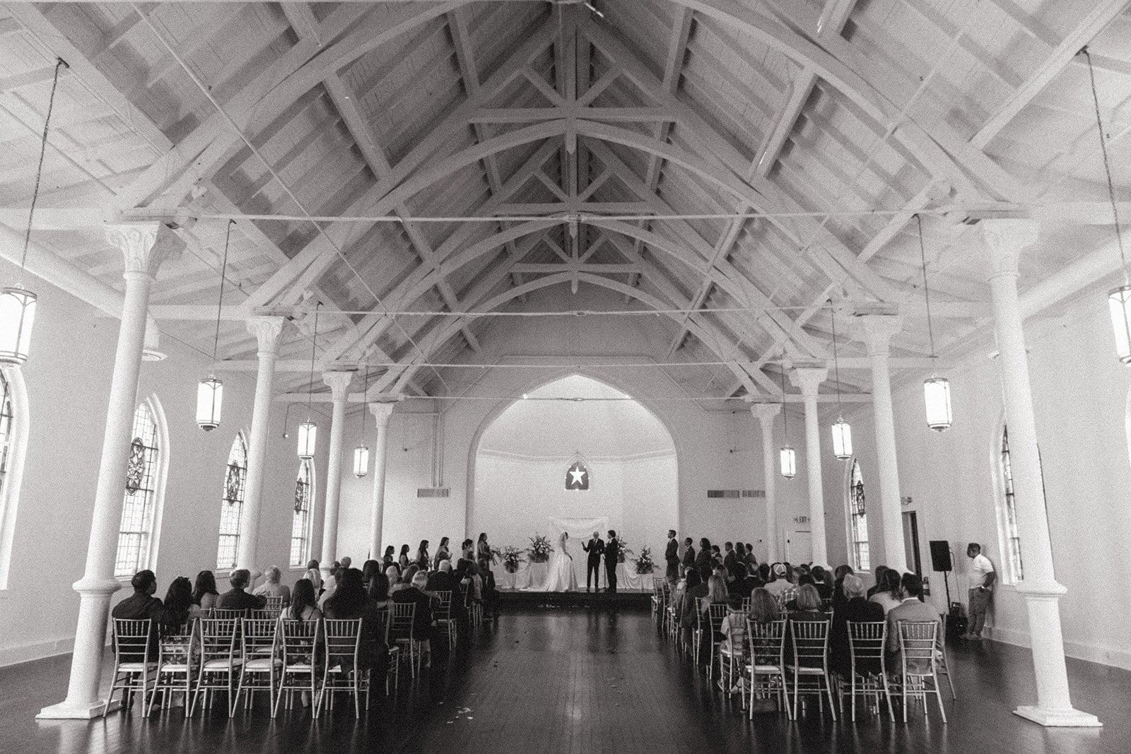 A wedding ceremony taking place inside a spacious hall with high vaulted roof, white columns, and large windows. The bride and groom stand at the front under a star-shaped decoration, surrounded by guests seated in chairs.
