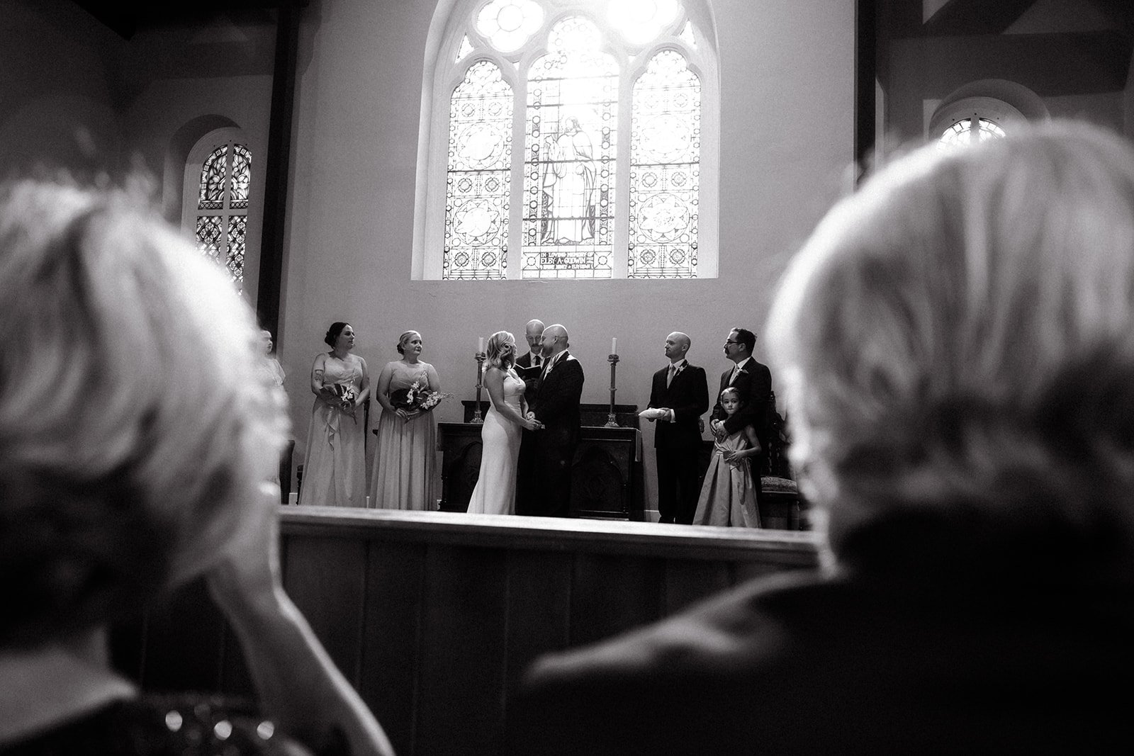 A black and white photo of a wedding ceremony inside a church, with the bride and groom holding hands and facing each other, surrounded by bridesmaids and groomsmen, and a large stained glass window in the background.