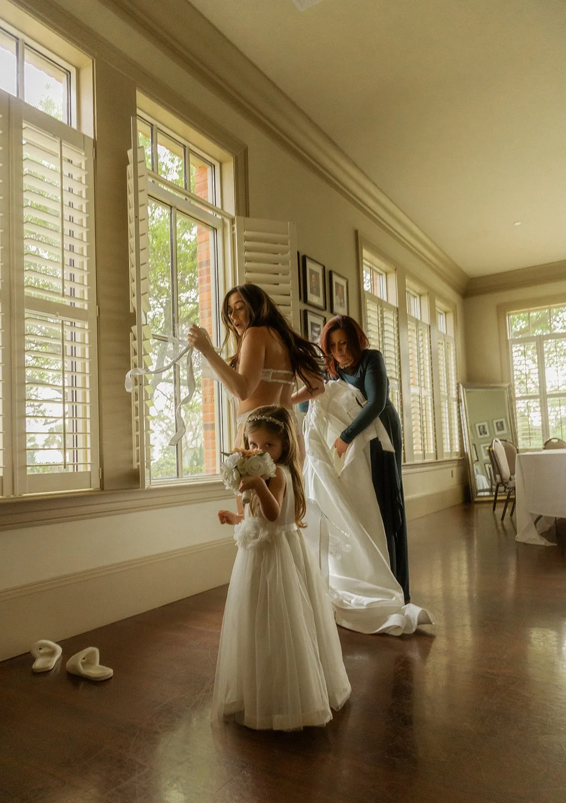 A woman in a wedding dress and a girl in a white dress holding a bouquet of white roses are standing by a window, with another woman helping the bride with her gown. Shoes are on the floor, and the room has multiple large windows with shutters and fr