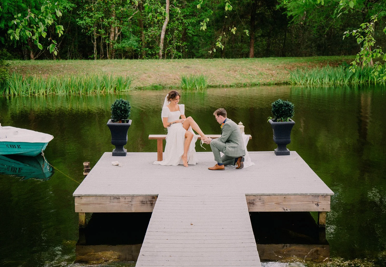 A couple getting married on a wooden dock by a river, surrounded by greenery and trees, with a boat to the left in the water.