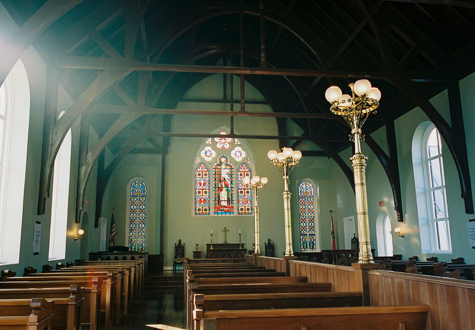 Interior of a church with wooden pews, stained glass windows, chandeliers, and a decorated altar at the front.