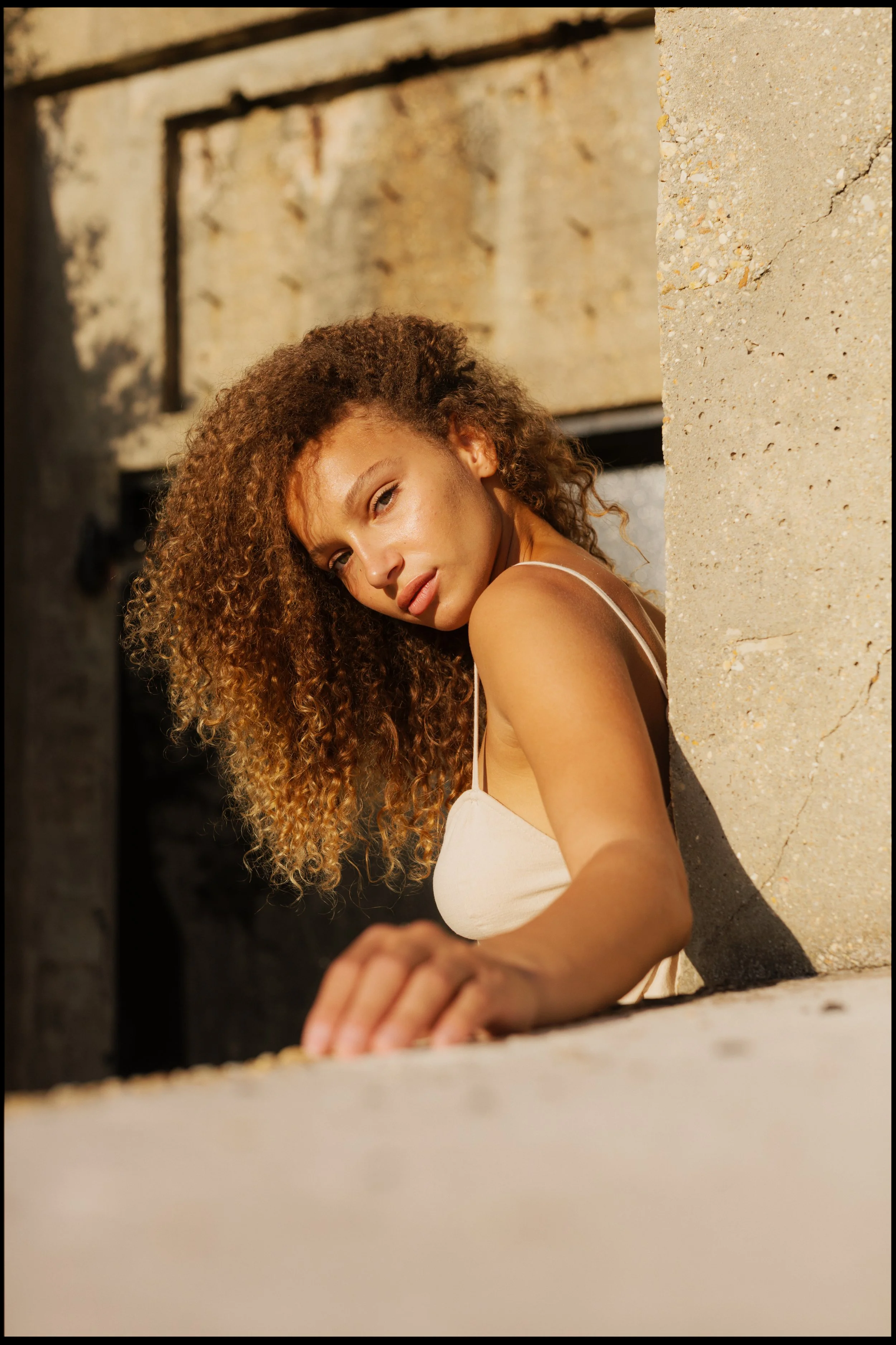 A woman with curly hair and a light-colored dress leaning on a concrete ledge, looking towards the camera with sunlight on her face, against an urban background.