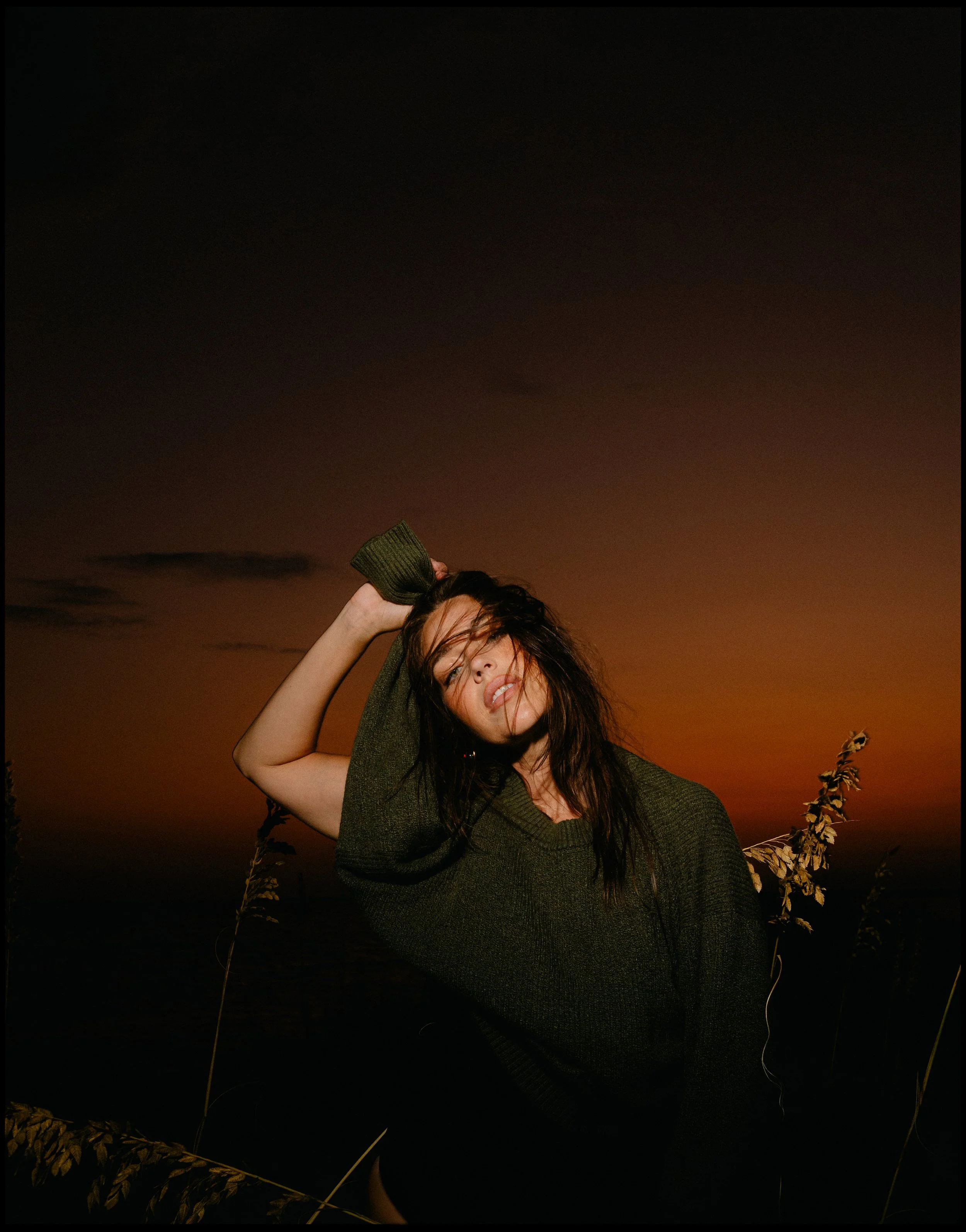 A woman with long dark hair, wearing a dark green sweater, posing outdoors during sunset with her hand behind her head and hair blowing in the wind.