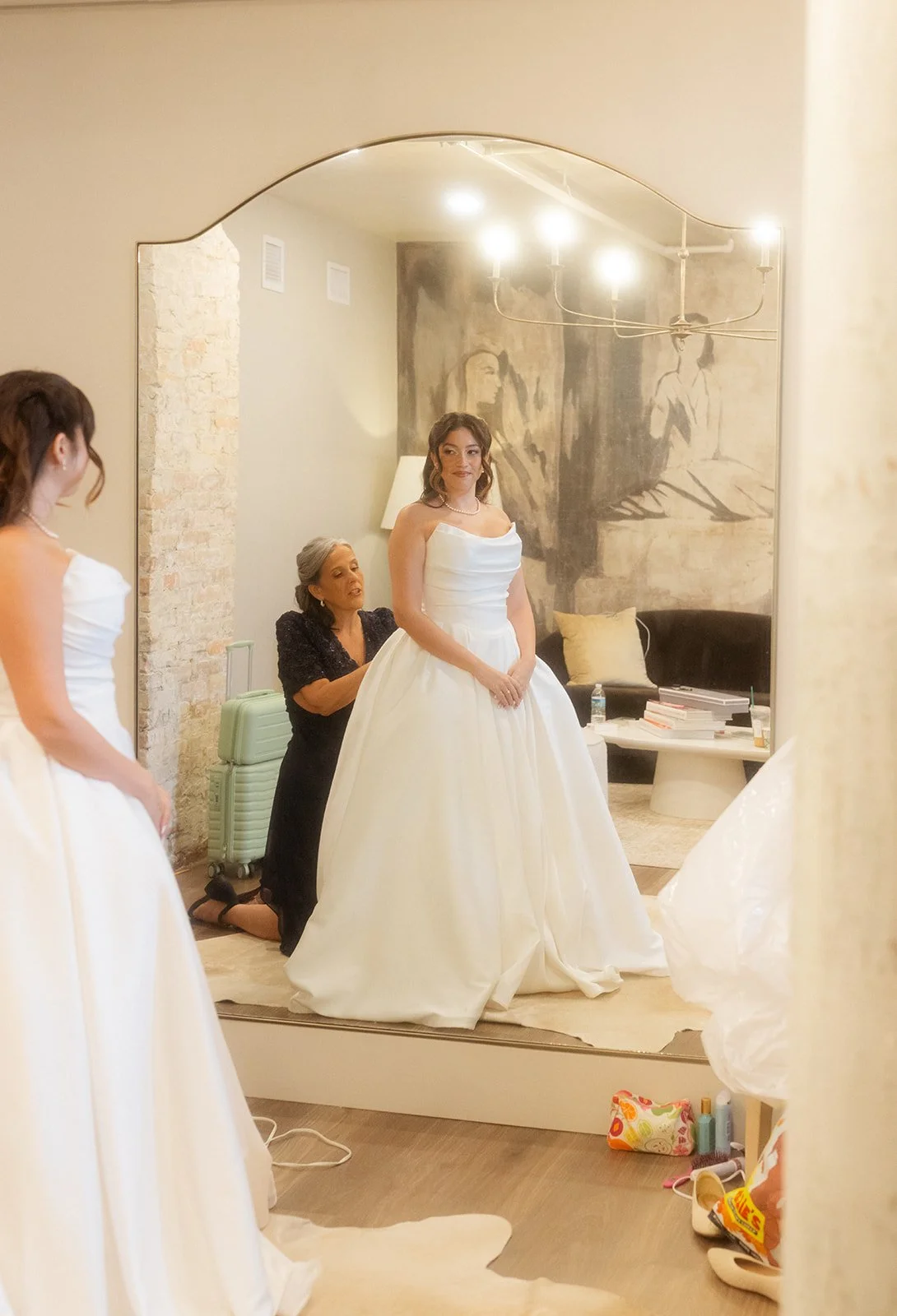 A woman in a wedding dress looks at herself in a mirror while a woman on her knees adjusts her dress, with other women nearby, in a room with artwork on the wall and a chandelier above.