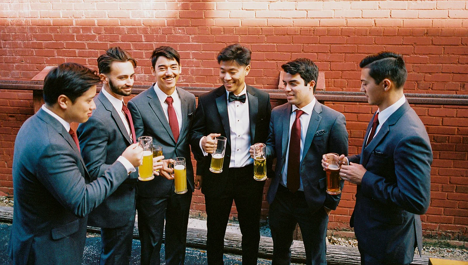 Group of seven men in suits holding drinks at a social gathering against a brick wall.