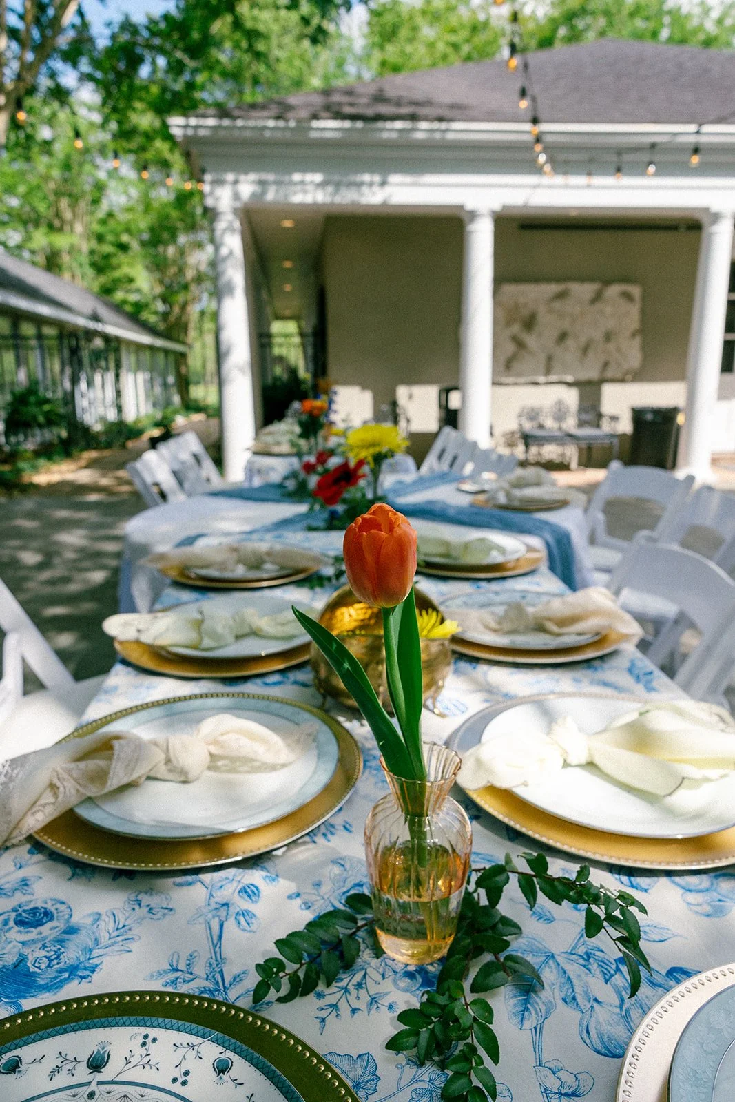 Outdoor dining table set with colorful flowers in vases, white plates with napkins, and a blue floral tablecloth, under string lights with a house in the background.