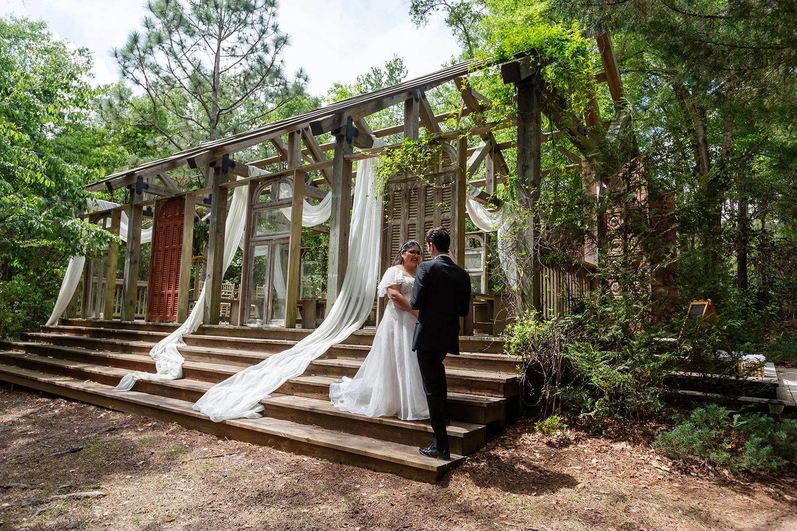 A bride and groom exchanging vows outdoors on a wooden platform with greenery around, under a rustic wooden arch decorated with fabric and plants.