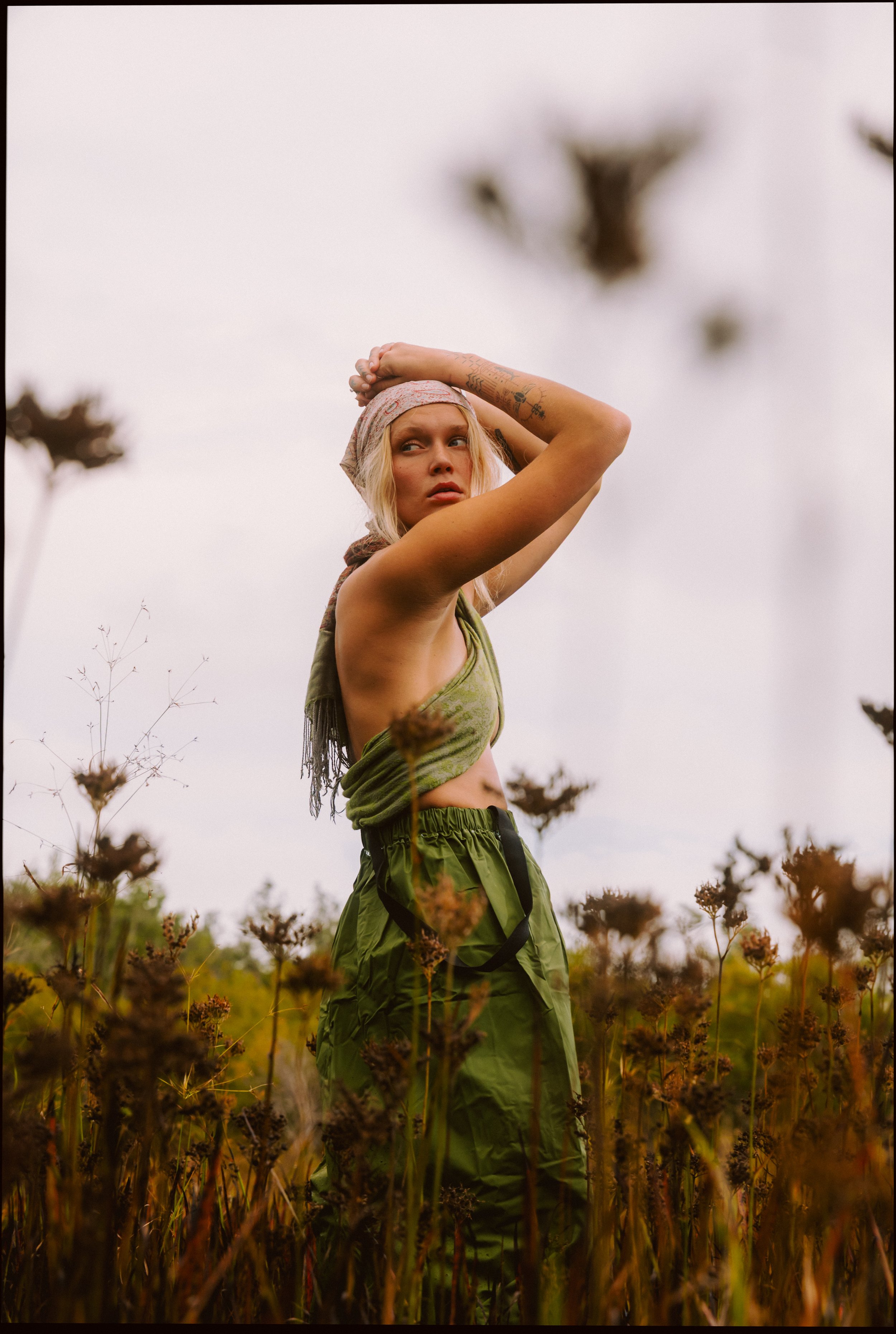 A woman standing in a field of dry plants, looking to the side with her arms raised above her head, wearing a bandana, green tank top, and green pants.