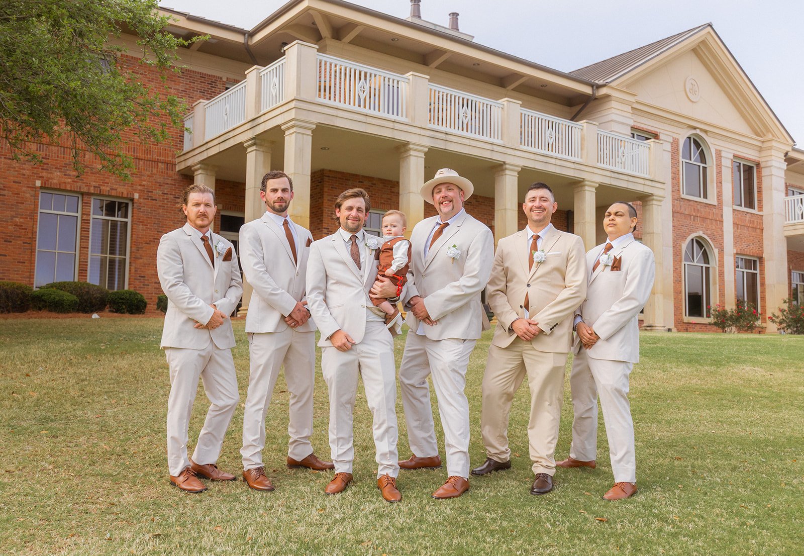 Group of men in beige suits and white pants standing on grass in front of a large brick house with columns and balconies, posing for wedding photo with one child held by a man.