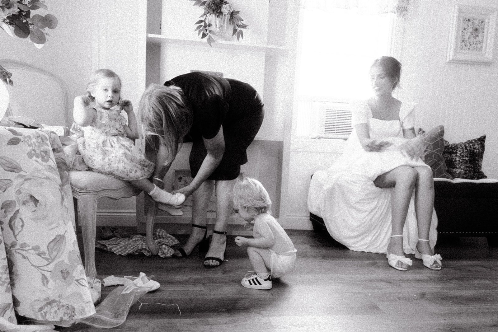 Two women and two young girls in a living room with floral couch and pillows. One woman is helping a girl put on shoes, while the other woman sits smiling on a sofa.