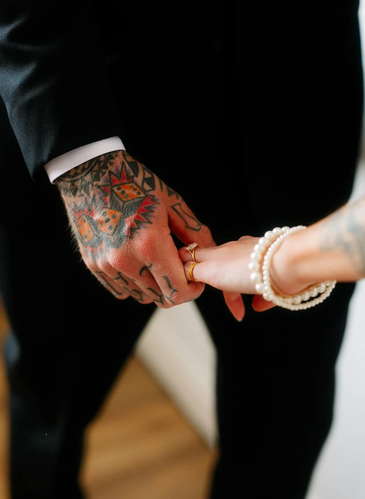 Close-up of a man and woman holding hands; the man has tattoos and wears a suit, the woman wears pearl bracelets and rings.