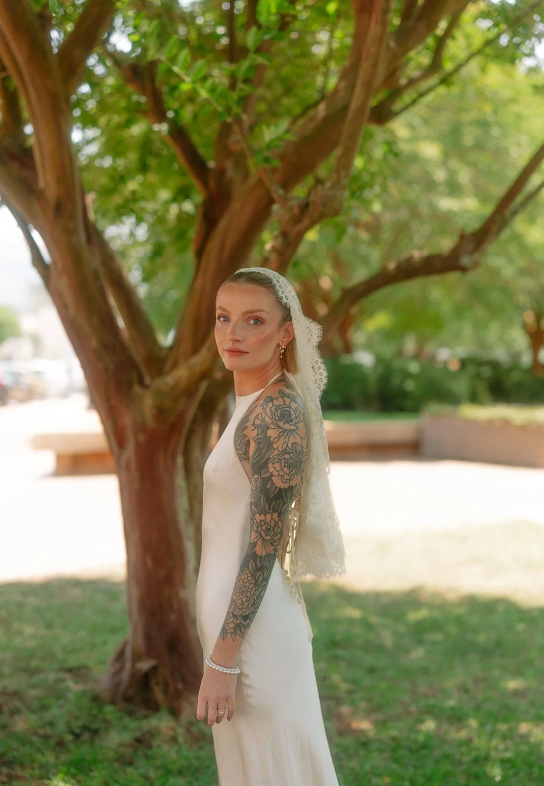 A woman in a white dress and lace veil standing outdoors near a large tree with green foliage, with visible tattoos on her left arm, in sunlight.