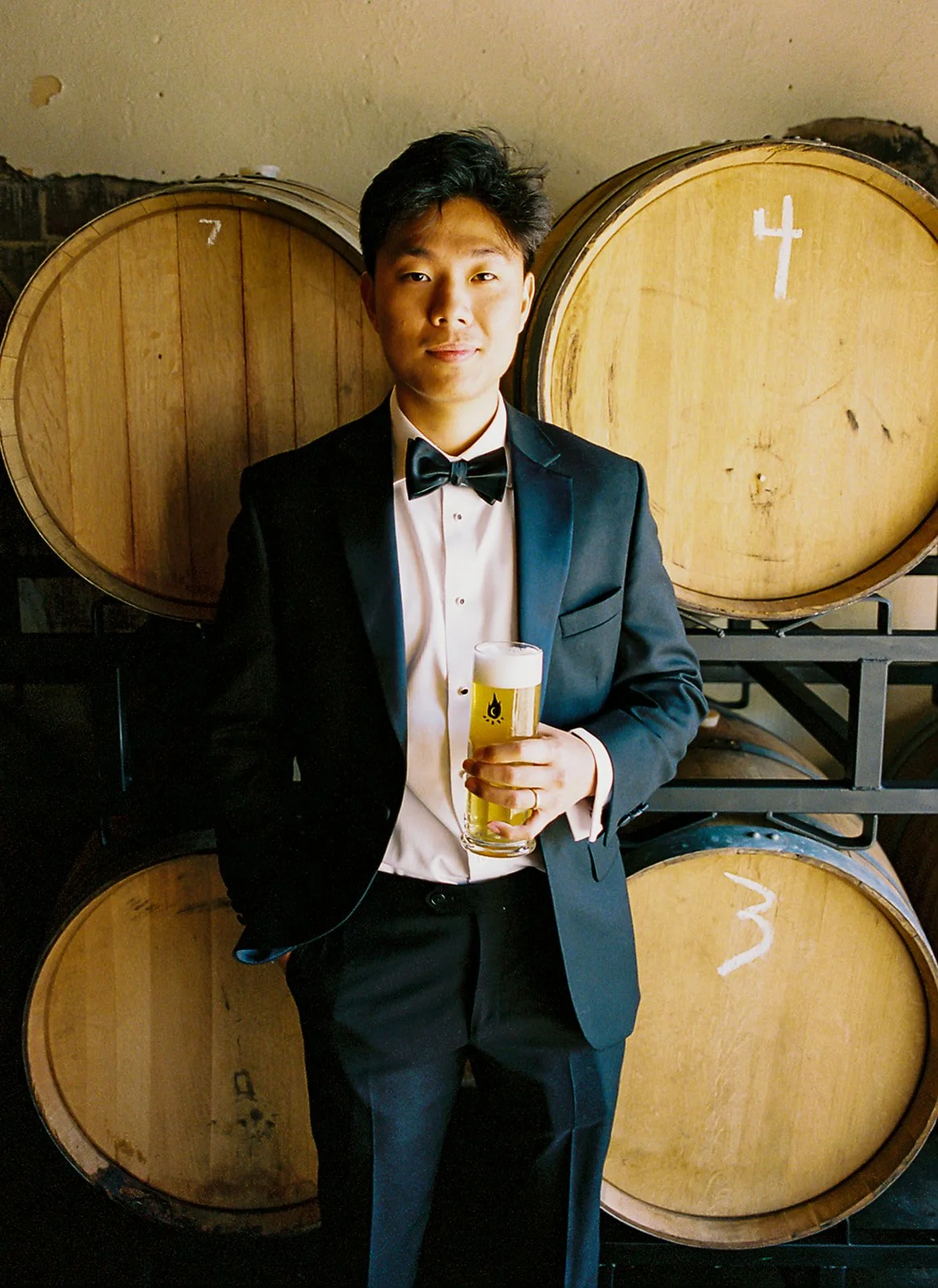 A young man in a tuxedo holding a glass of beer, standing in front of large wooden barrels labeled with the numbers 4 and 3.