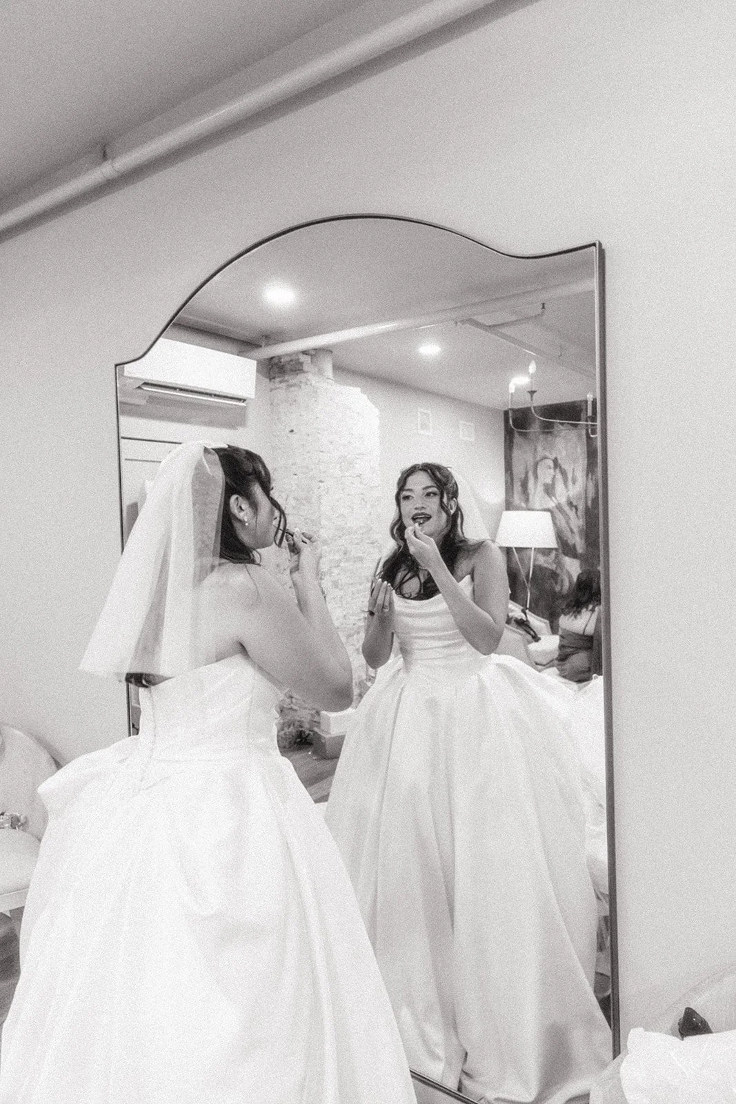 Two women in wedding dresses applying makeup while looking at each other in a mirror.