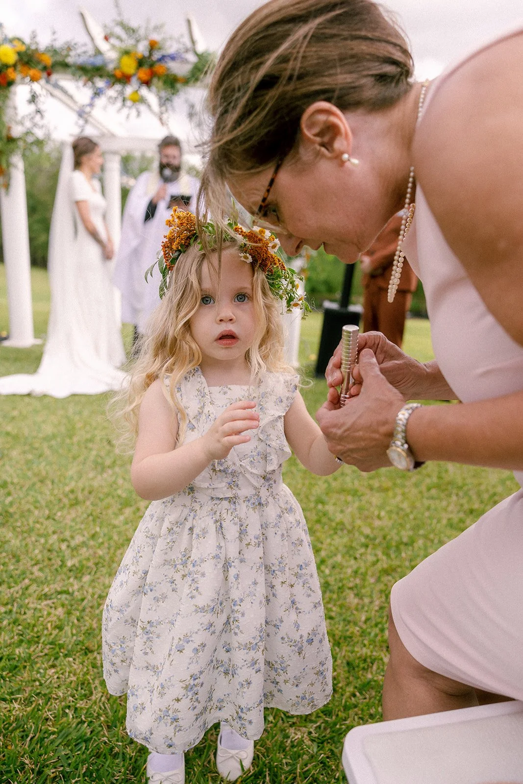 A young girl with curly blonde hair, wearing a floral dress and a flower crown, stands outdoors during a wedding ceremony. An older woman, possibly the officiant, bends towards her, holding a metallic object. In the background, a couple stands under 