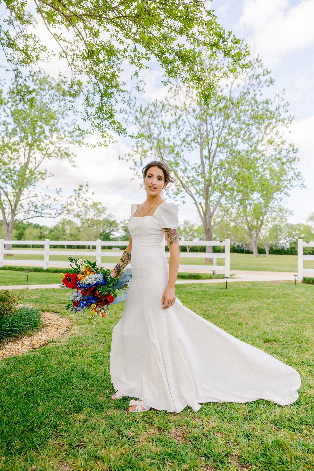 A bride in a white wedding dress holding a colorful bouquet outdoors on a grassy lawn with trees and a white fence in the background.