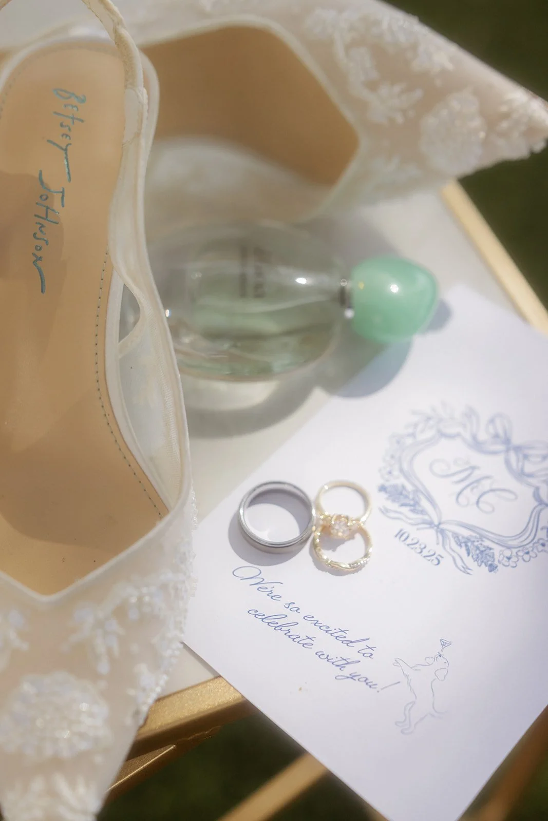 Wedding invitation with rings, a glass perfume bottle with a green cap, and a wedding shoe with pearl embellishments.