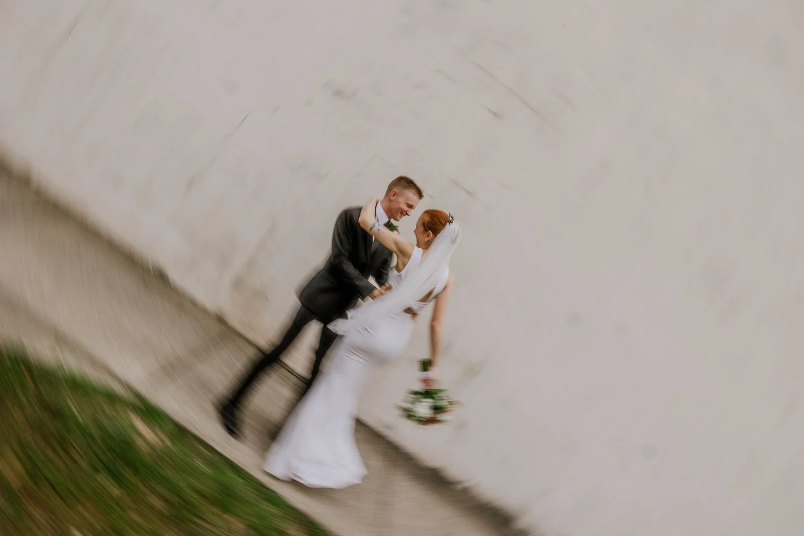 A newlywed couple dancing outside against a white wall, with the bride holding a bouquet, both smiling and looking at each other.