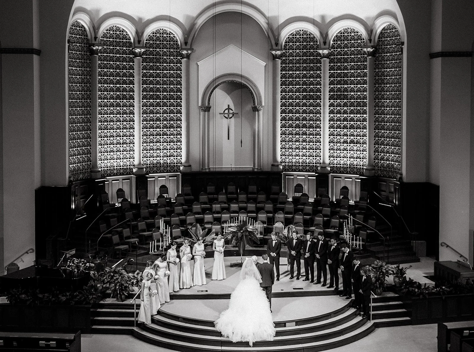 A black and white photo of a wedding ceremony taking place in a church. The bride and groom stand at the altar, surrounded by bridesmaids and groomsmen, with the church's large stained glass windows and high altar in the background.