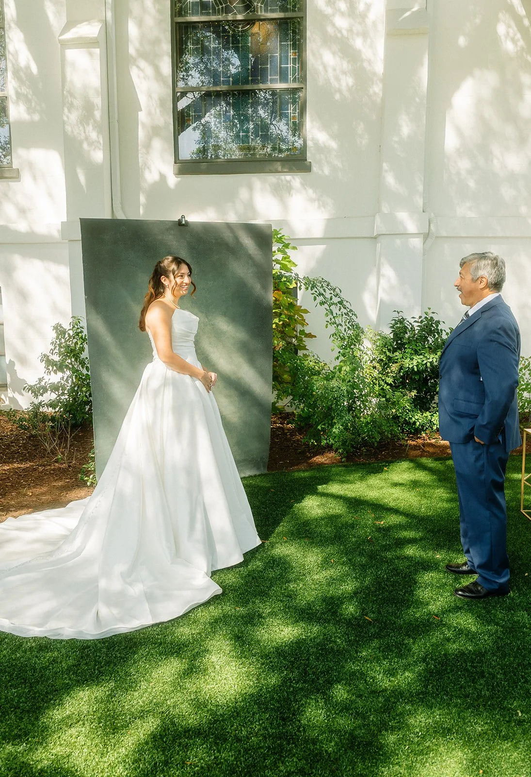 A bride and an older man in a suit are standing outside on a grassy area, facing each other. The bride is wearing a white wedding gown and smiling, while the older man is smiling back. There's a dark backdrop behind the bride and some green bushes ar