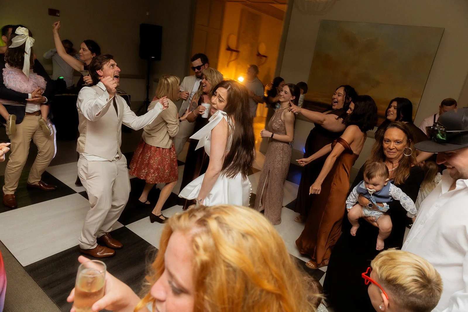 People dancing and celebrating at a party, with some holding drinks and children present, in an indoor venue with a checkered dance floor.