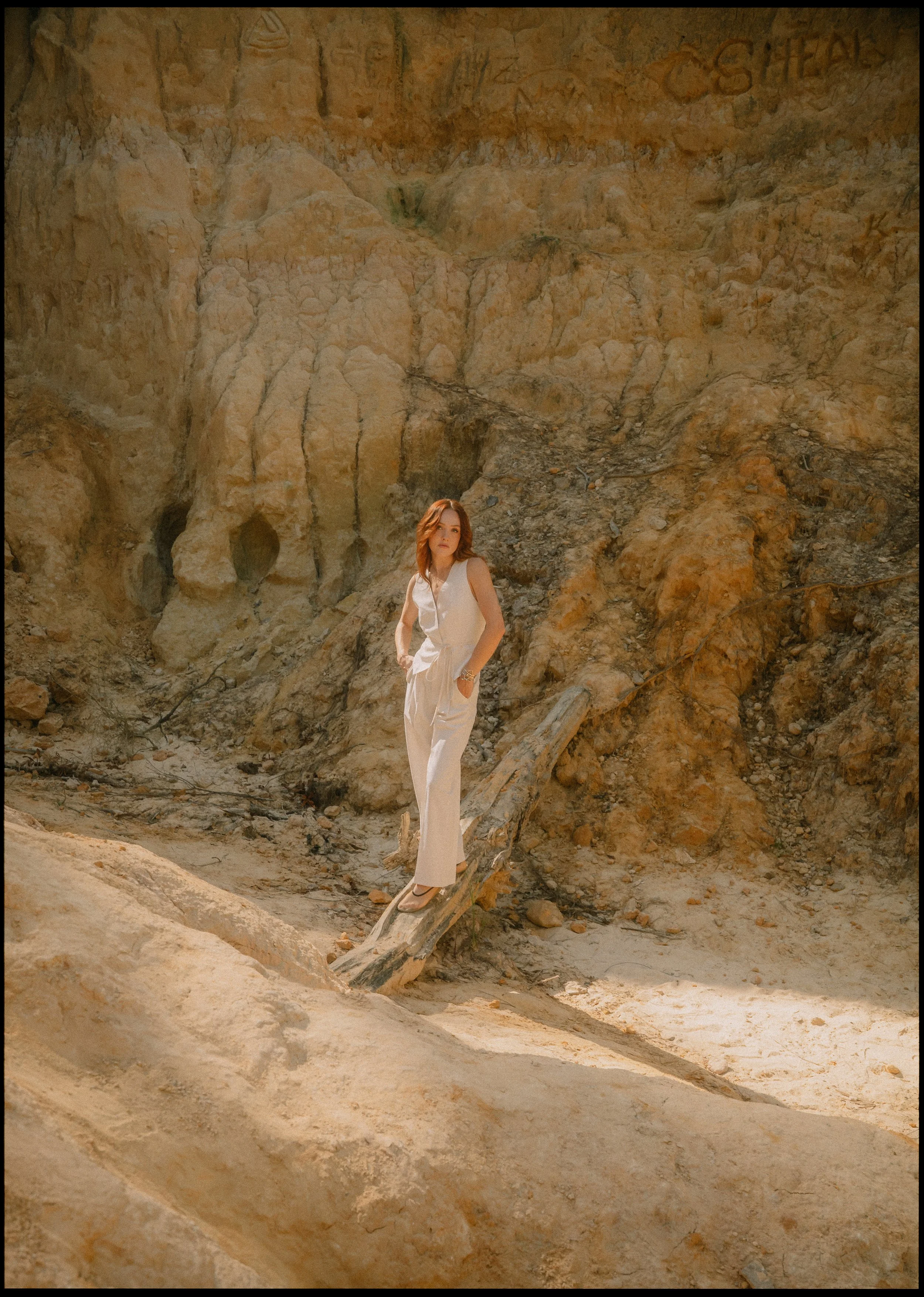 A woman in a white jumpsuit standing on a fallen tree trunk in a desert landscape with rocky cliffs in the background.
