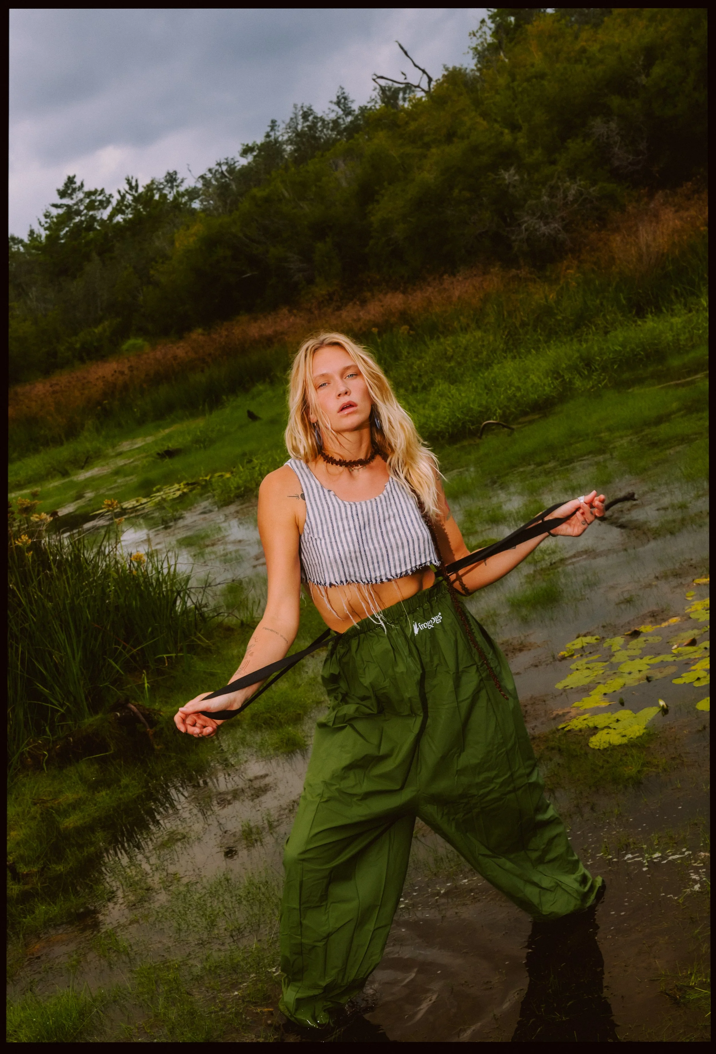 A young woman standing in a wetland area with water and lily pads, surrounded by lush green vegetation and trees, under a cloudy sky.