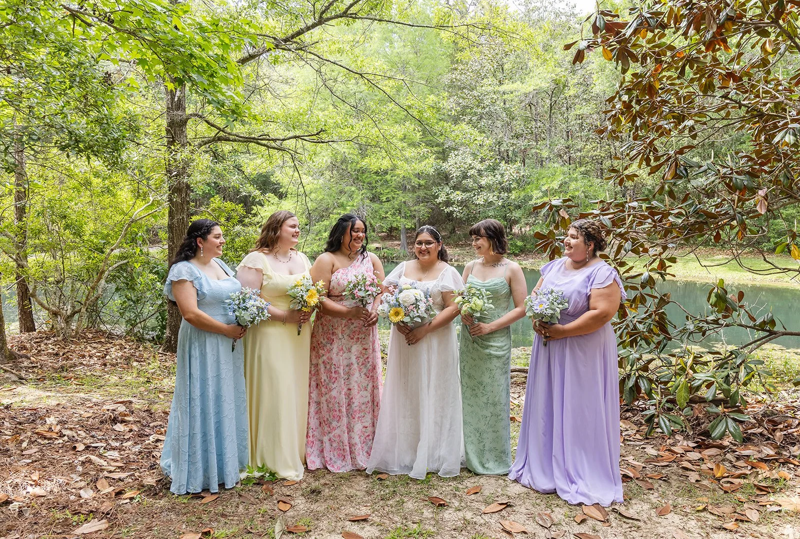 Six women in colorful dresses holding bouquets of flowers, standing on a wooded area near a lake, smiling and talking to each other during daytime.