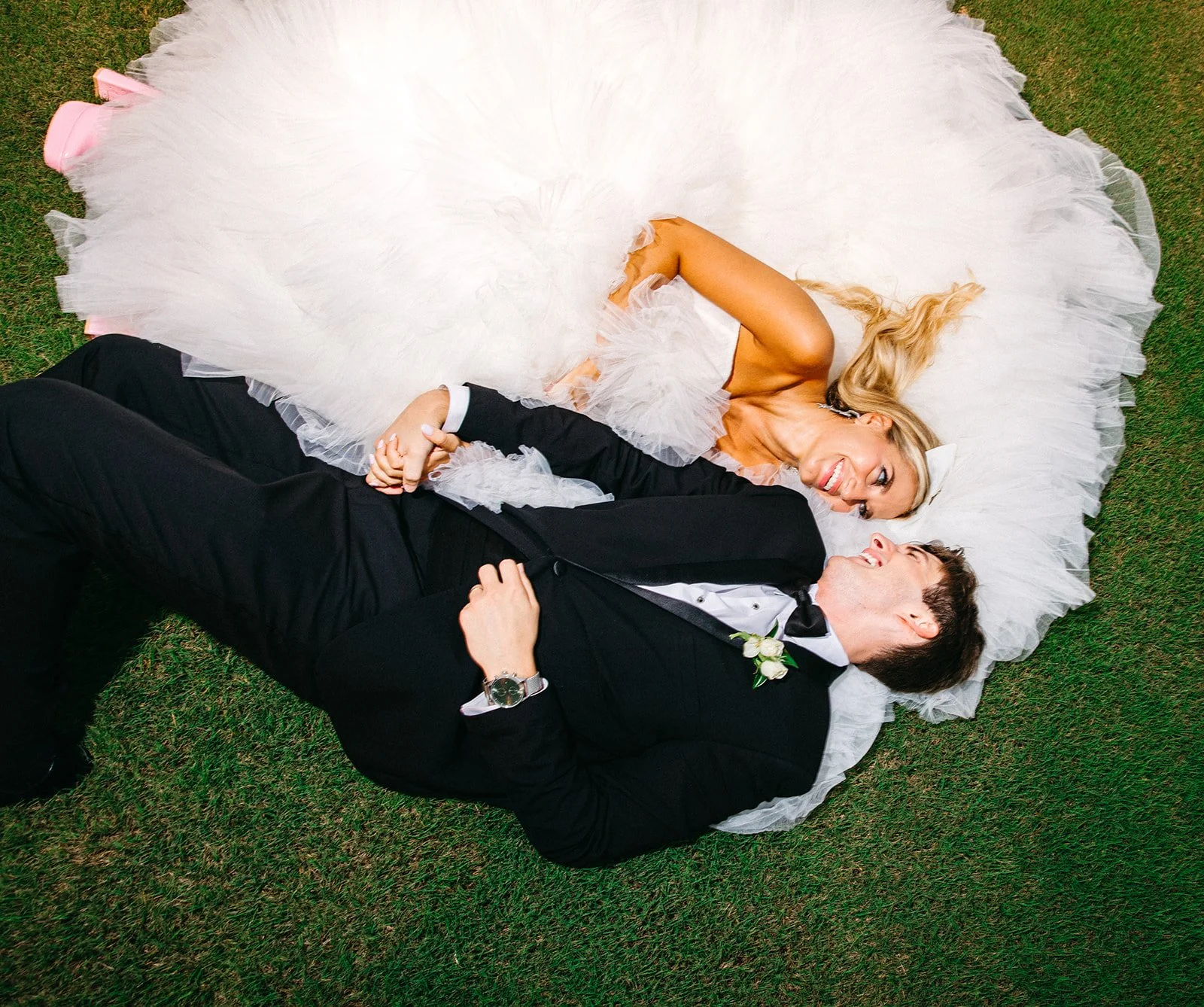 A bride and groom lying on the grass, holding hands, smiling at each other. The bride is wearing a white wedding dress with a voluminous skirt, and the groom is dressed in a black tuxedo with a boutonniere.