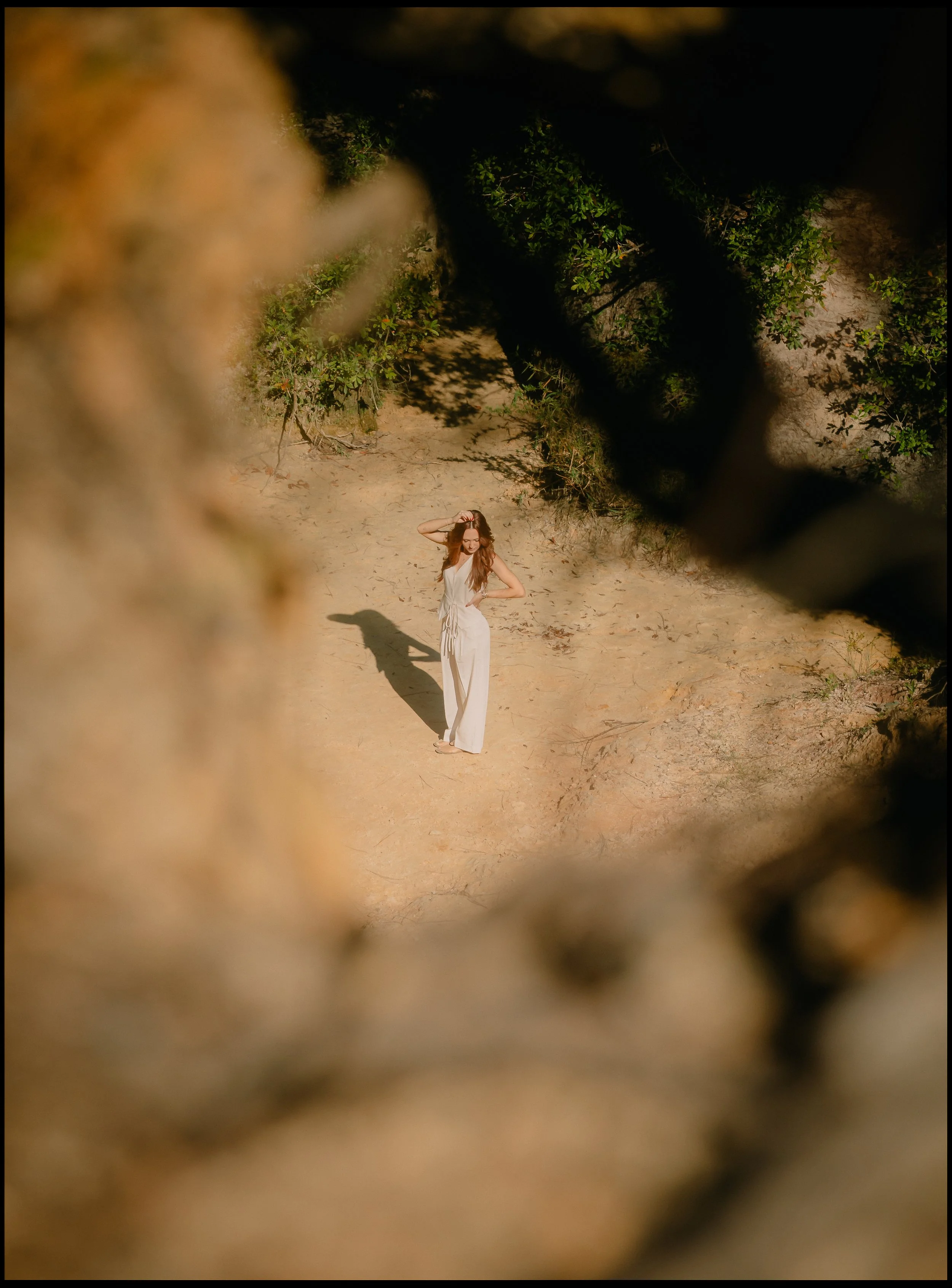 A woman in a white dress standing on a sandy area, viewed through a blurry chain-link fence with foliage nearby.