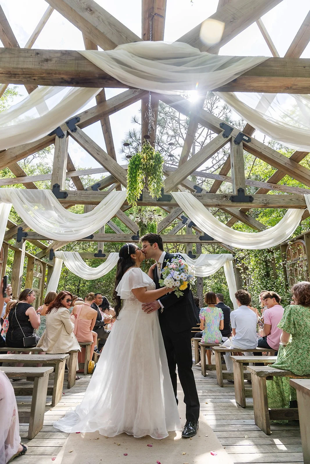 A bride and groom kiss during their outdoor wedding ceremony under a wooden pergola decorated with white fabric and greenery, with guests seated on benches watching.