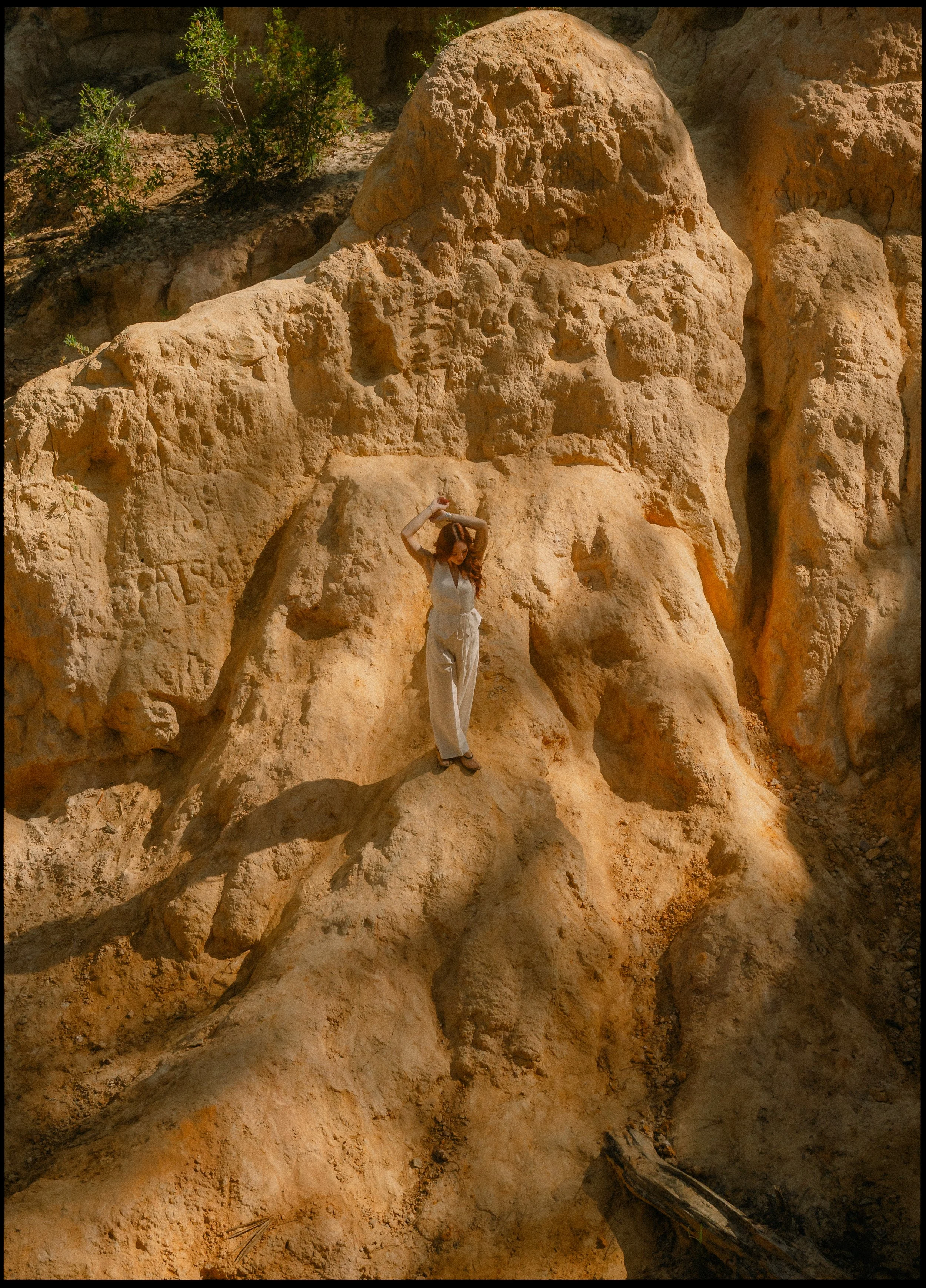 A woman standing on a large, rugged rock formation in a desert landscape, holding a hat overhead, with some green bushes at the top of the rocks and a shadow cast on the rocks.