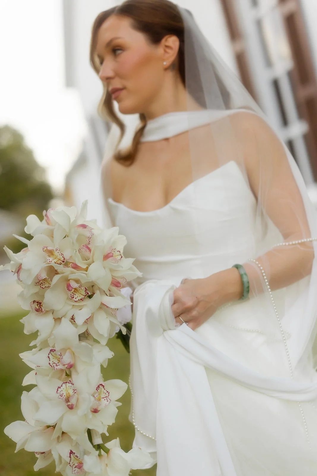 A bride in a white wedding gown holding a bouquet of white orchids, with a veil, jewelry, and a bracelet, standing outdoors near a building.