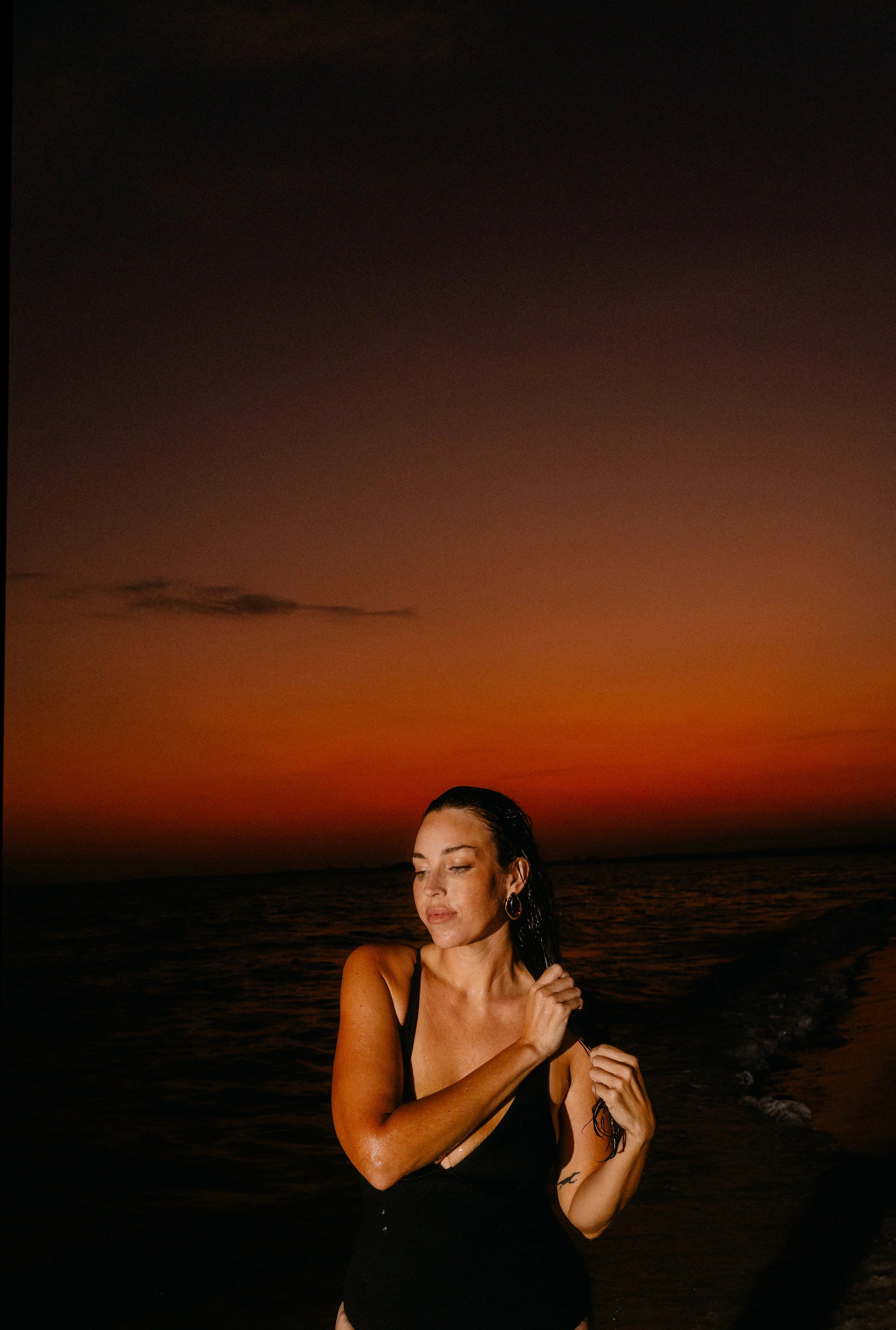 A woman in a black swimsuit wades in the ocean at sunset, with a colorful sky in the background.