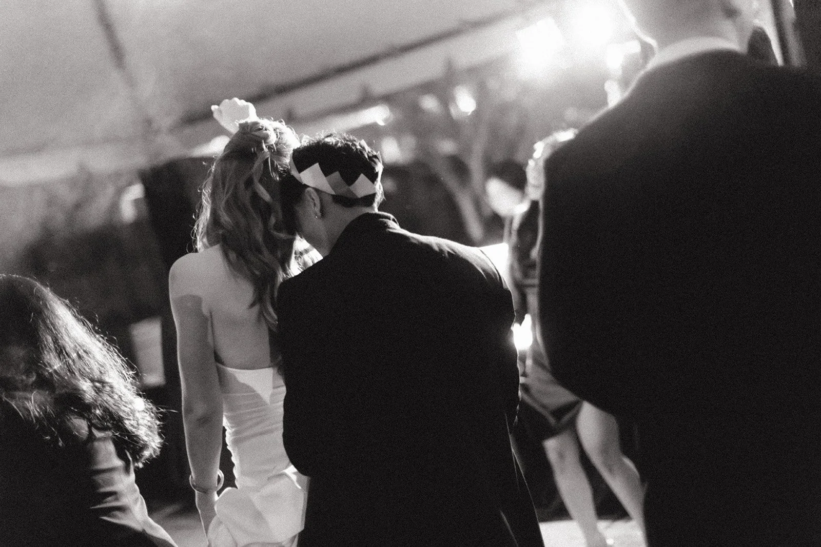 A black and white photo of a couple kissing at a party or event, with other people in the background, and warm lighting.