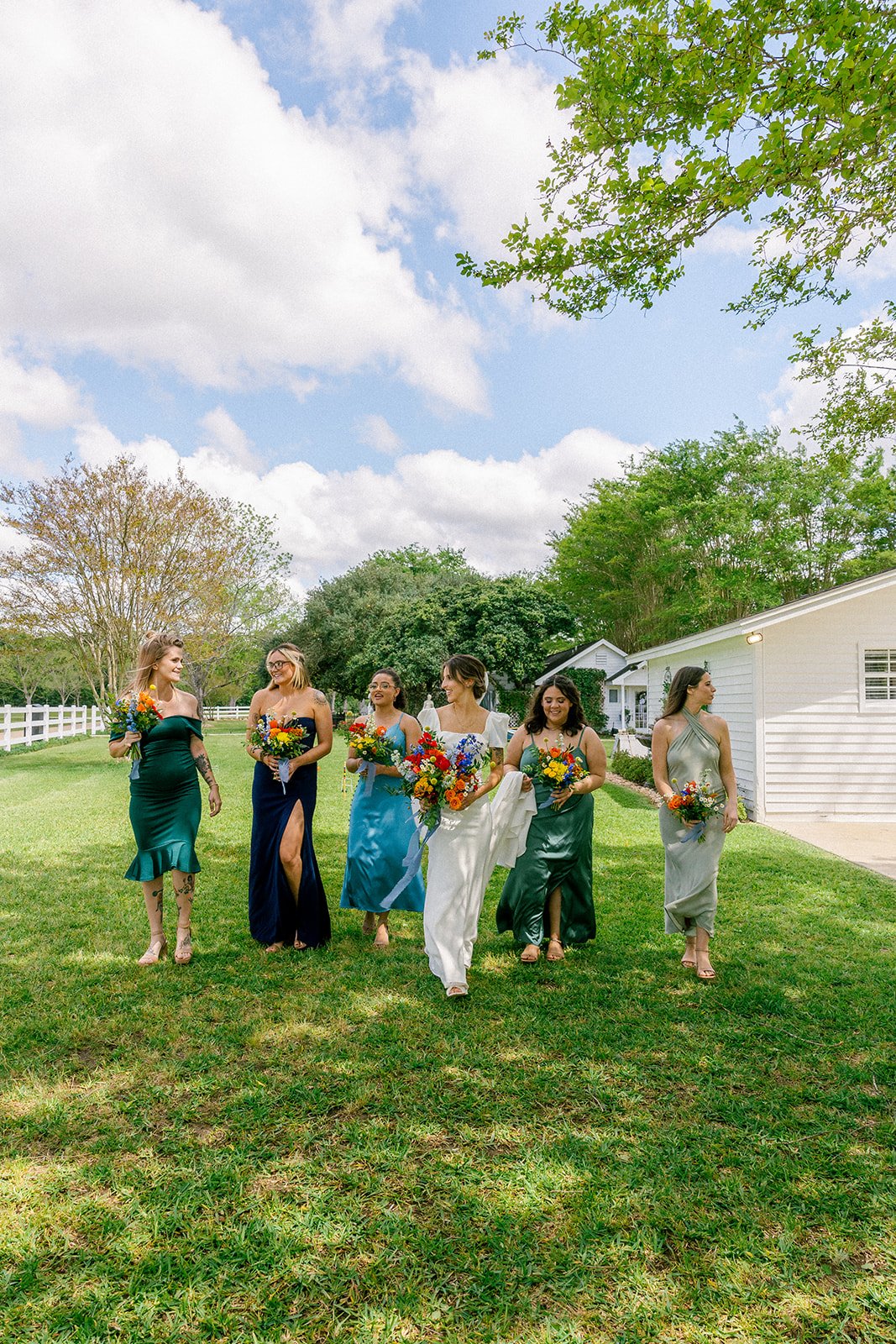 Group of women in colorful dresses holding bouquets walking on a grassy lawn outdoors, with trees and a white building in the background under a partly cloudy sky.